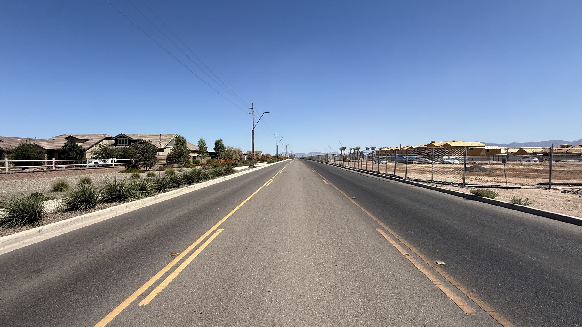 Wide desert street, clear blue sky. Left: single-family homes with fencing and landscaping; right: chain-link fence around a construction site. Utility poles line the road; mountains on the horizon.