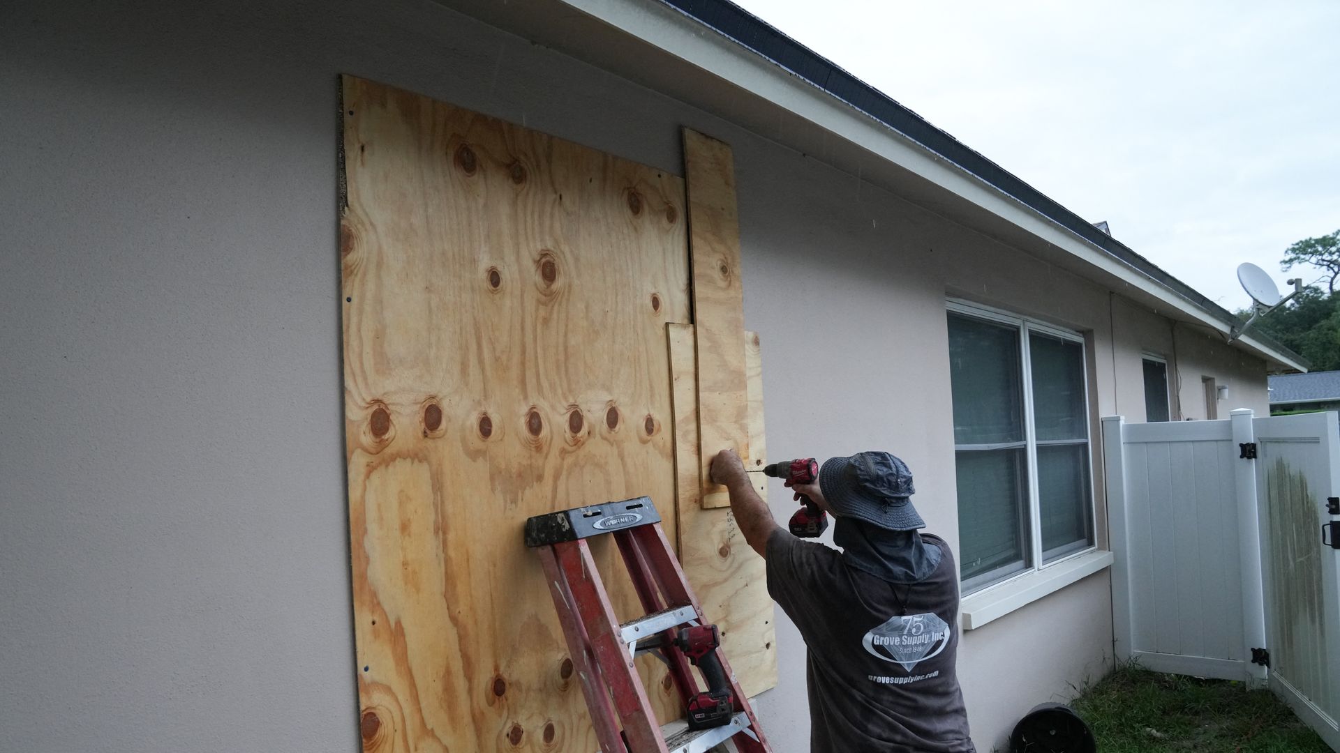 A man in a gray bucket hat drills plywood over windows of his home.