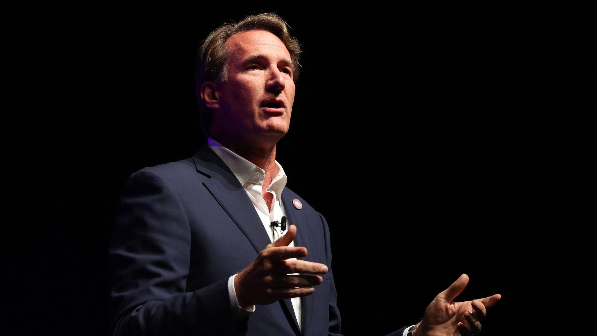 Gov. Youngkin in a suit speaking with his hands stretched out with a black background behind him