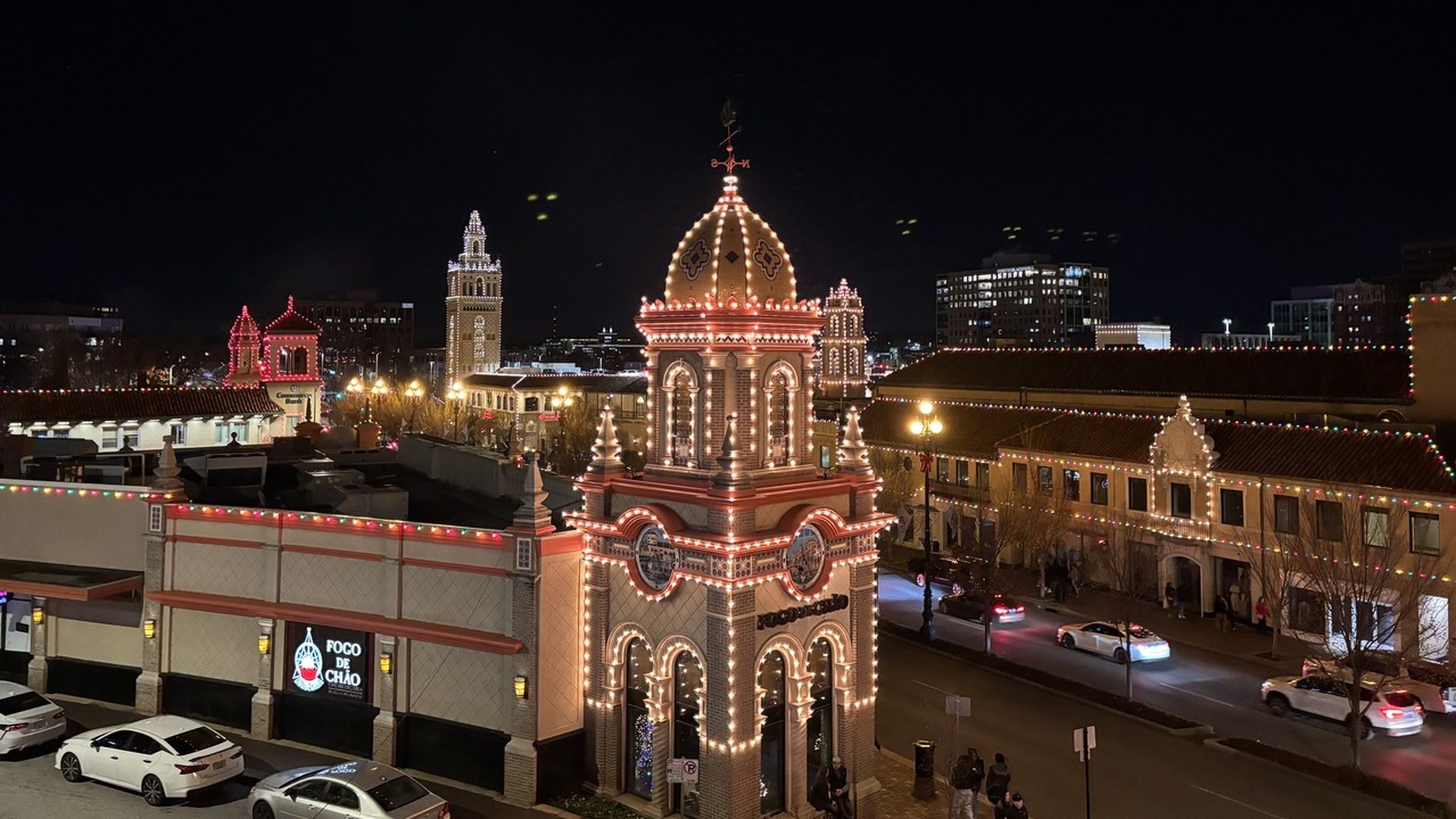 Night scene of a city street with historic buildings outlined in colorful holiday lights, including a prominent tower with warm white lights and red accents, and parked cars below.