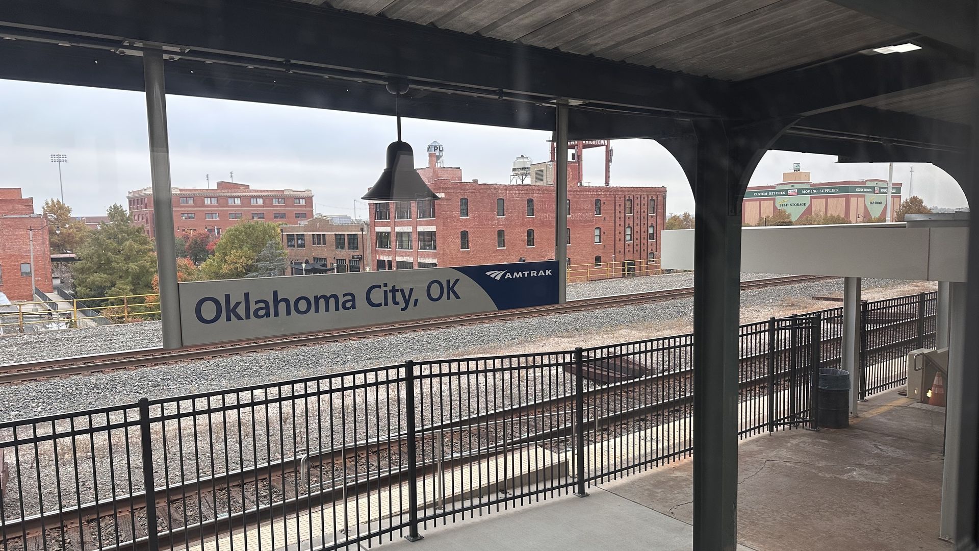 View of Amtrak train station platform in Oklahoma City, OK, with black railing, tracks, and brick buildings in background under a cloudy sky.