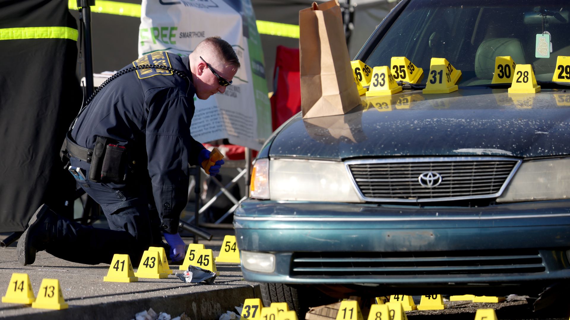 Investigator in dark uniform kneeling near a blue Toyota car covered with yellow numbered evidence markers on the ground and hood, investigating a crime scene in Oakland, Calif., with debris and a paper bag nearby.