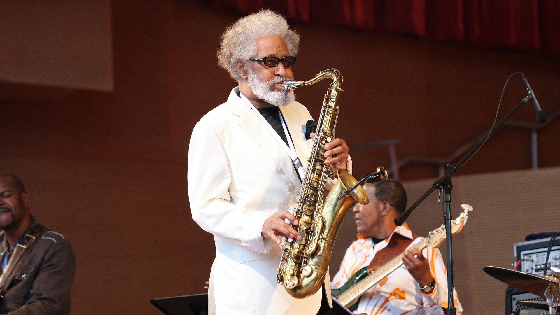 An older man with gray hair and beard, in a white suit and dark glasses, plays a saxophone on stage while red curtains rise behind and other musicians are visible.