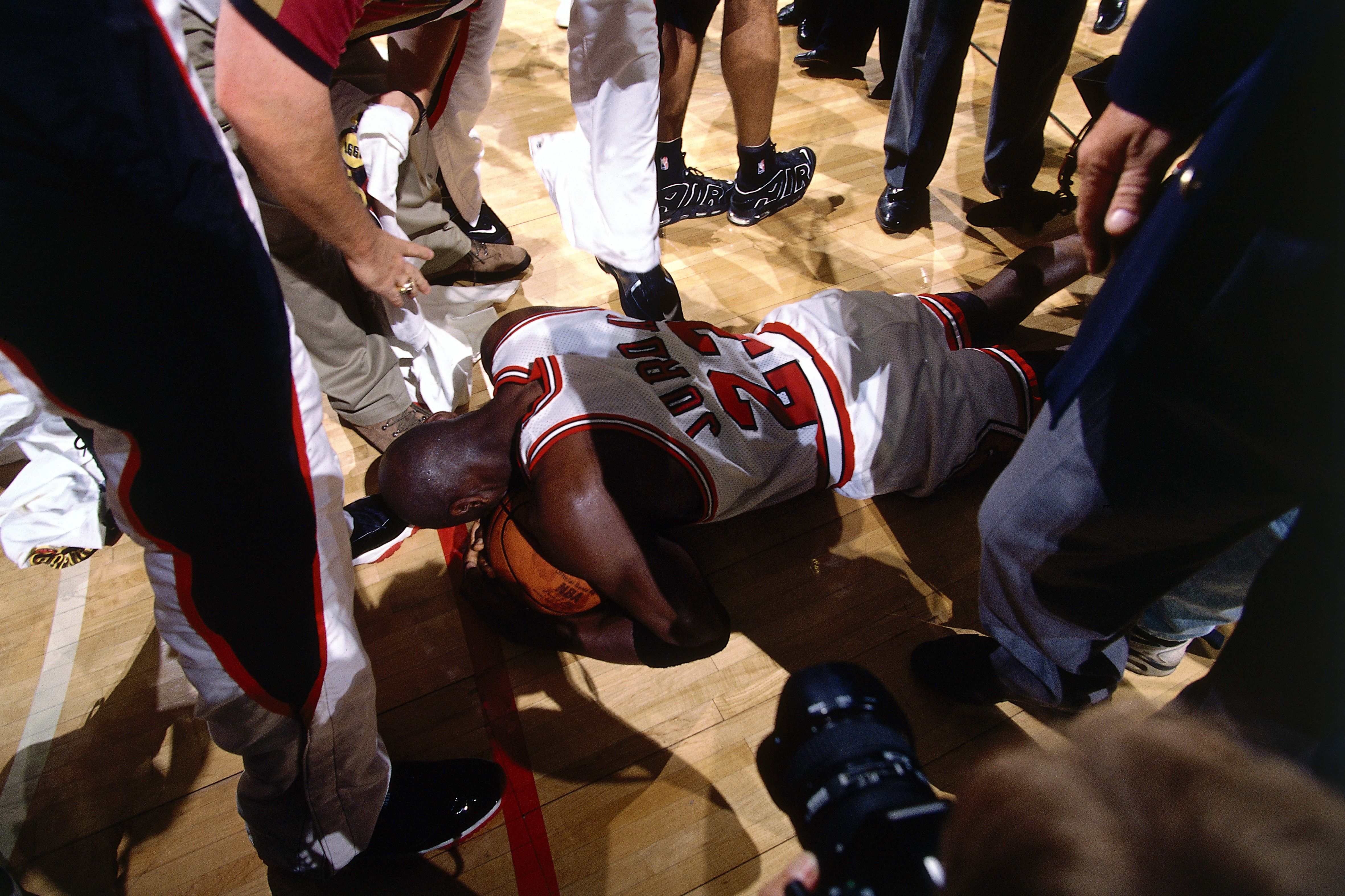 Basketball players crowd a fallen teammate on a polished wooden court; a white jersey with red trim lies on the floor while others stand or kneel around him in sneakers and uniforms.