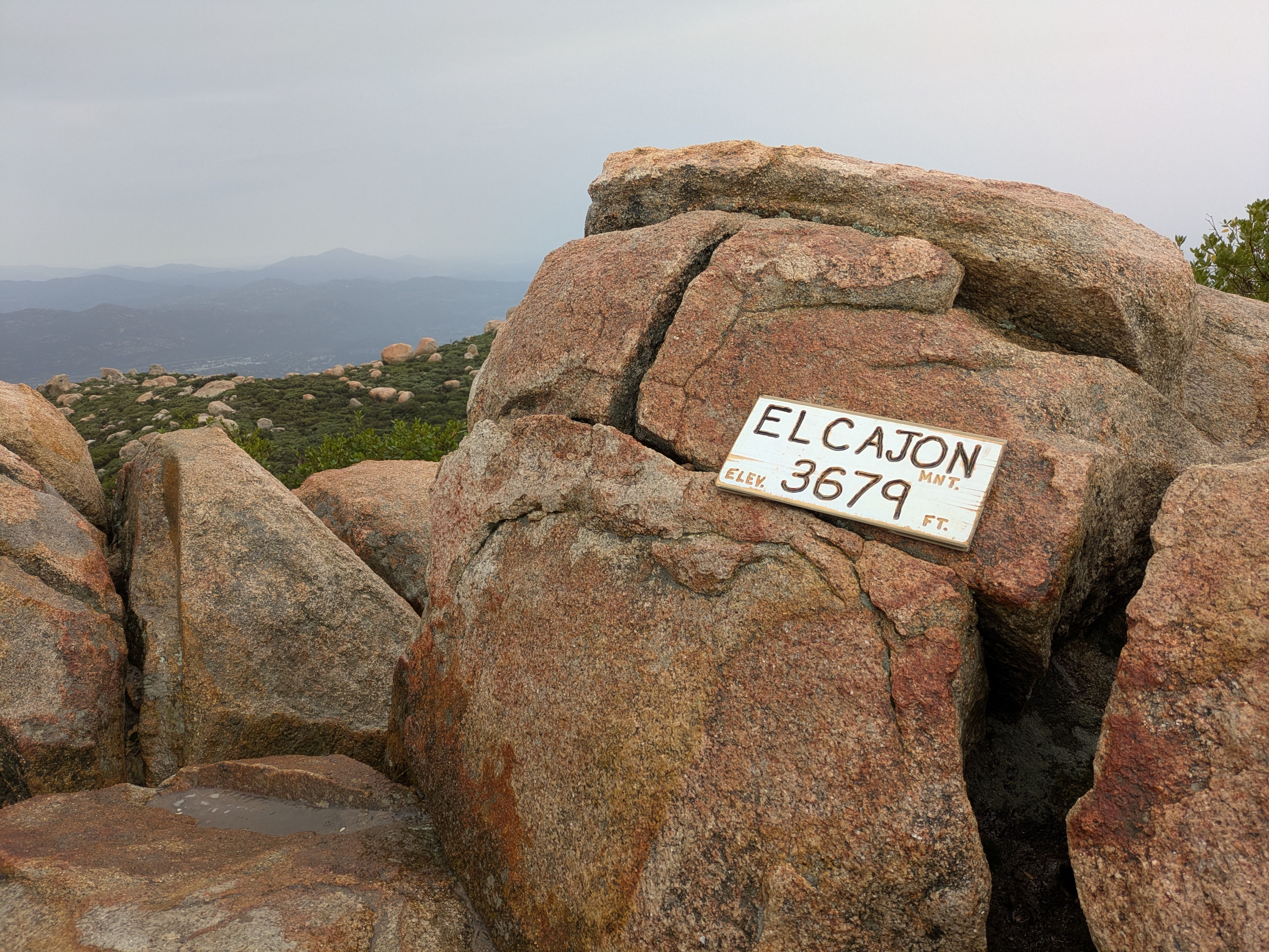 Large reddish-brown rocks on a mountain summit with a white sign reading "El Cajon Mnt. Elev 3679 Ft." Green shrubbery and distant hazy mountains under a gray sky in the background.