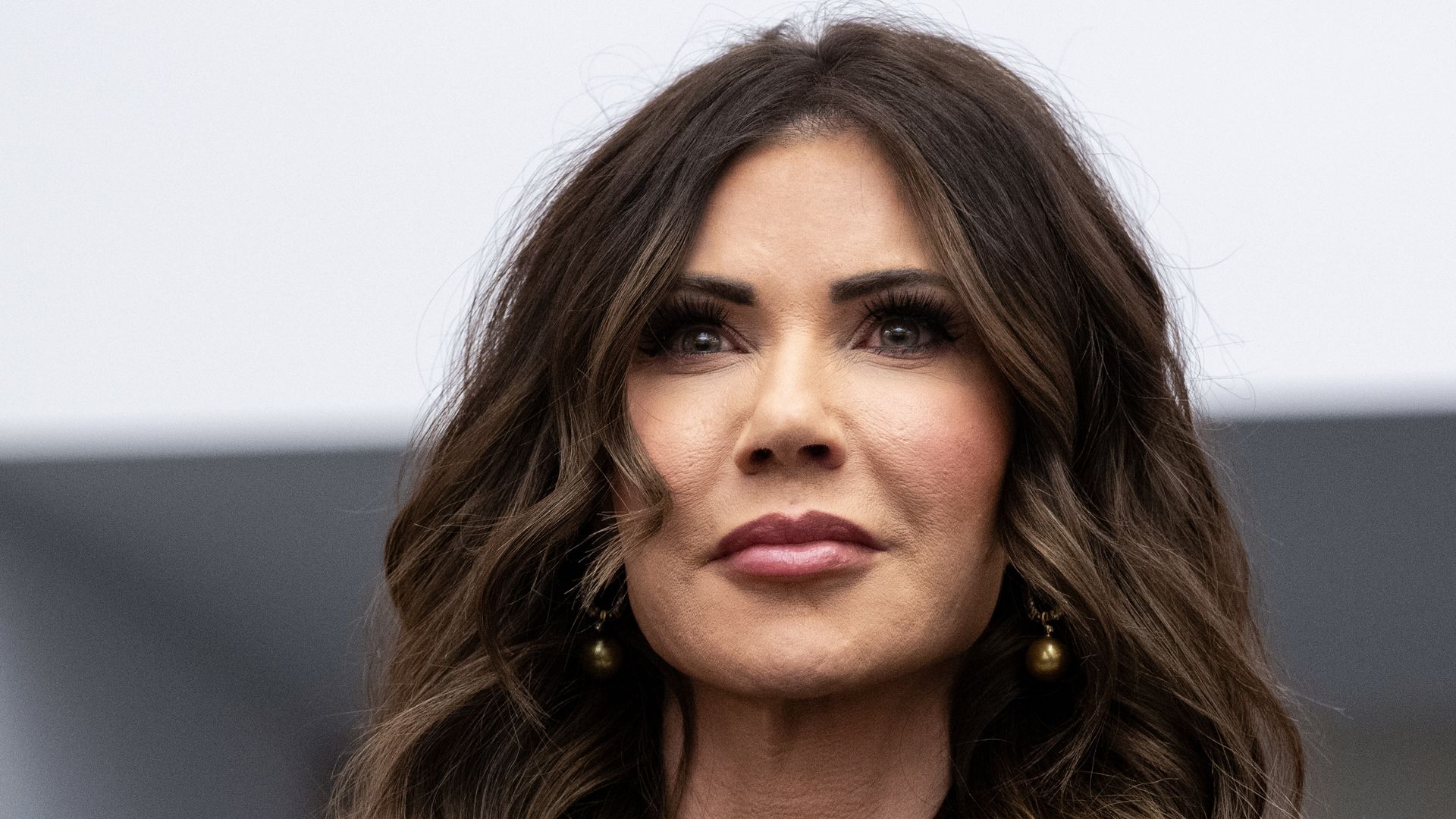 Close-up of a woman with long wavy brown hair, dark eyes and wearing dark round earrings against a blurry gray and white background.