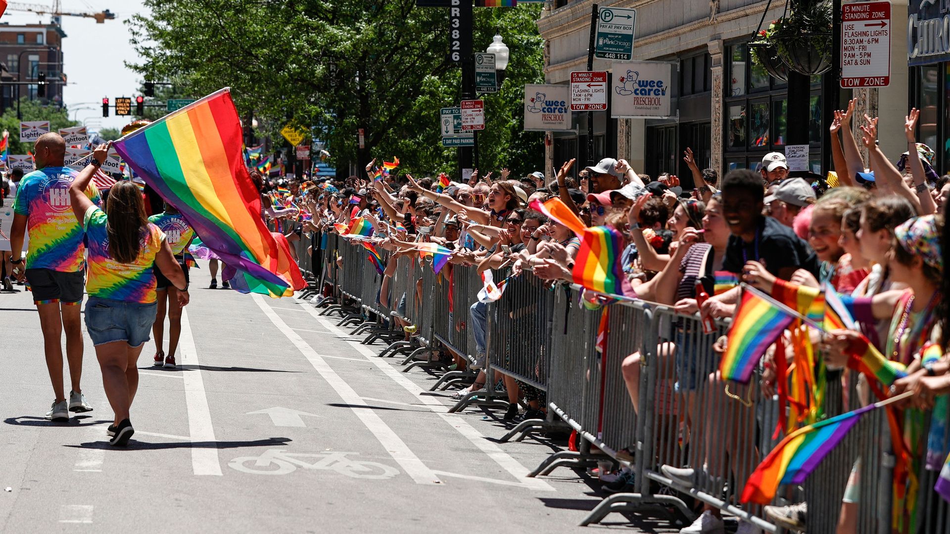 Crowd behind barriers along a city street, waving large rainbow Pride flags. Colorful shirts and banners line the right-side buildings while trees shade the left.