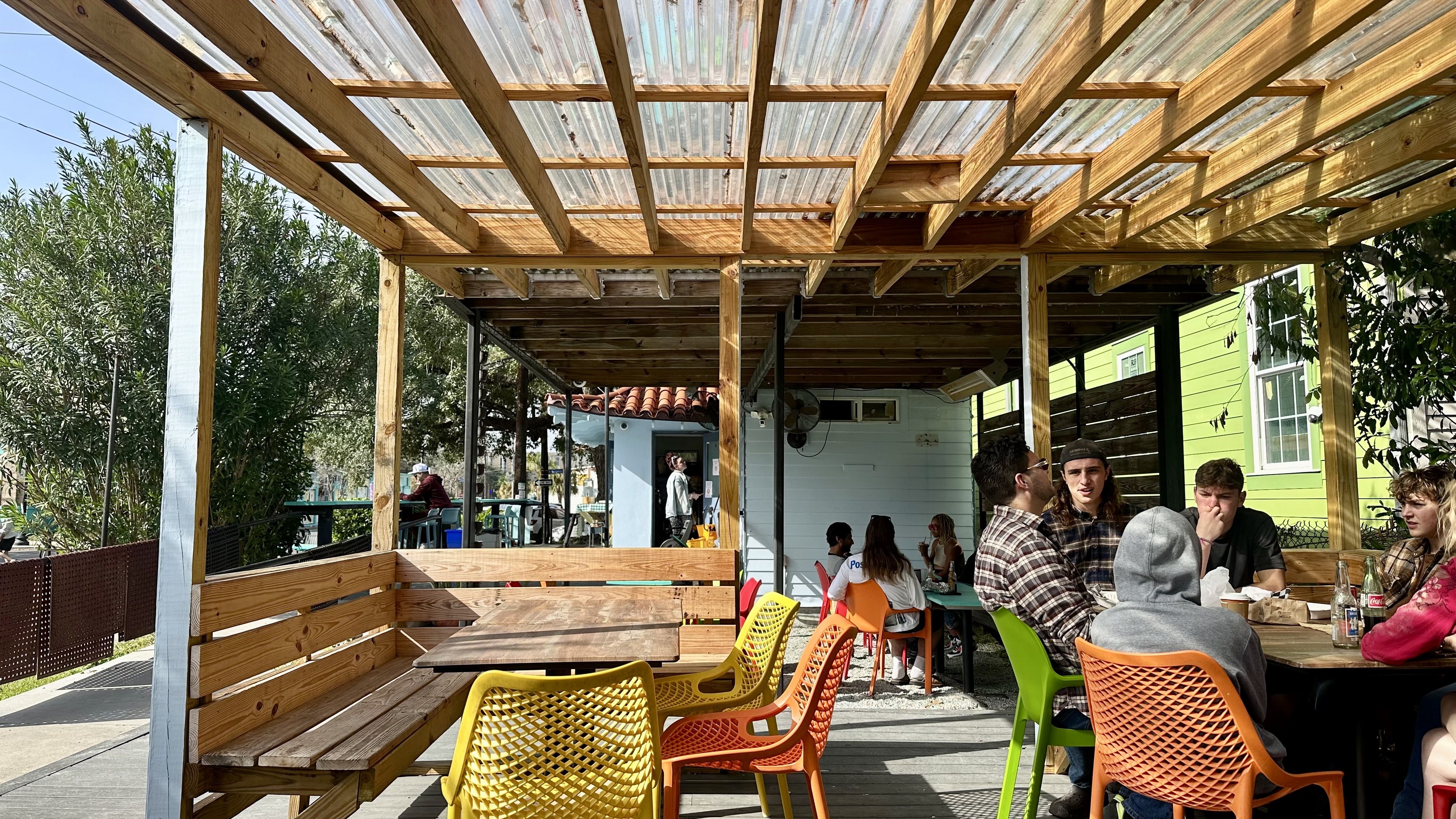Outdoor seating area with wooden benches and colorful plastic chairs (yellow, orange, green) under a clear corrugated roof. Several people sitting and talking around tables.