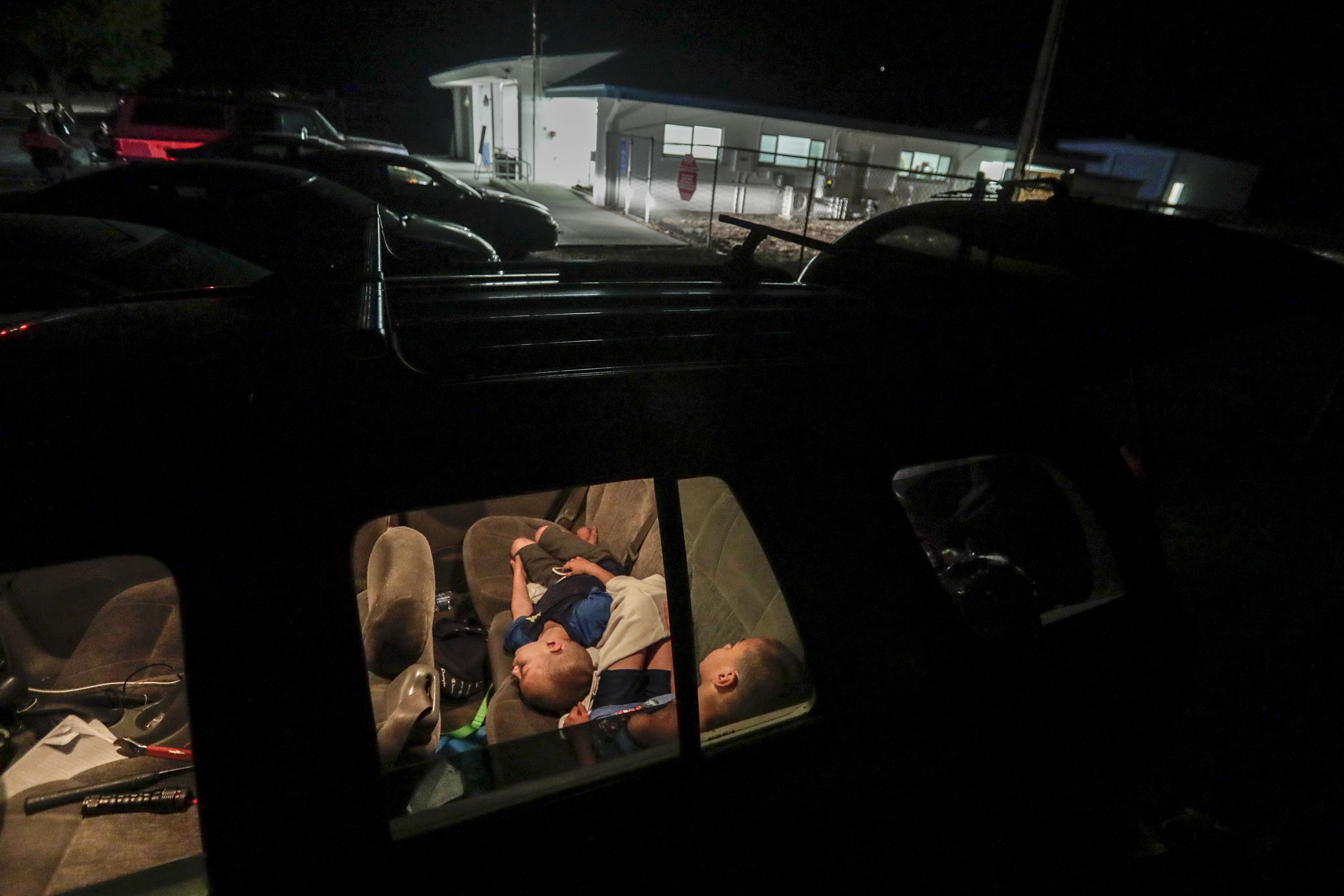This image shows infants and young children sleeping in the back of a car in a parking lot at night.