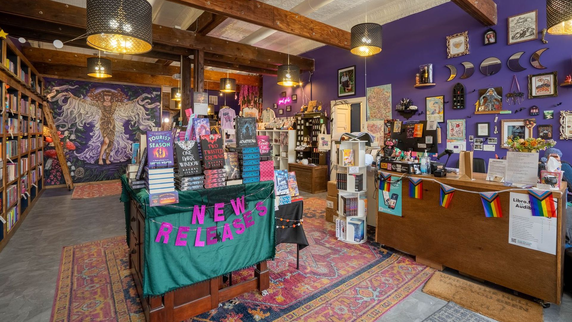 Cozy bookstore interior with purple walls, wooden beams, hanging black lamps, colorful rugs, a bulletin wall of framed art, and a central table displaying new book releases. 