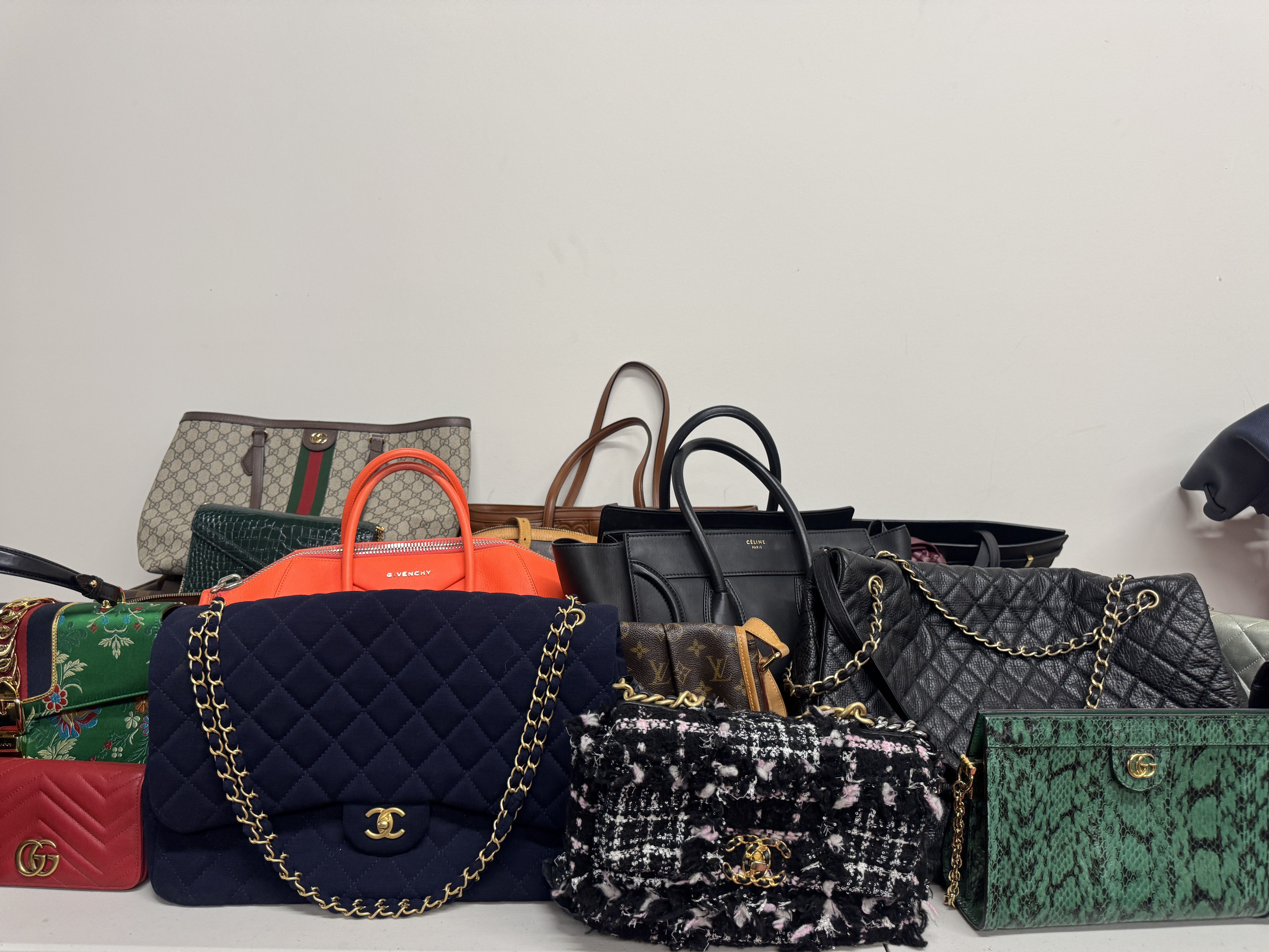 Collection of luxury handbags including black and navy quilted Chanel bags with gold chains, a green snakeskin Gucci bag, a bright orange Givenchy bag, and patterned and leather styles against a white wall.
