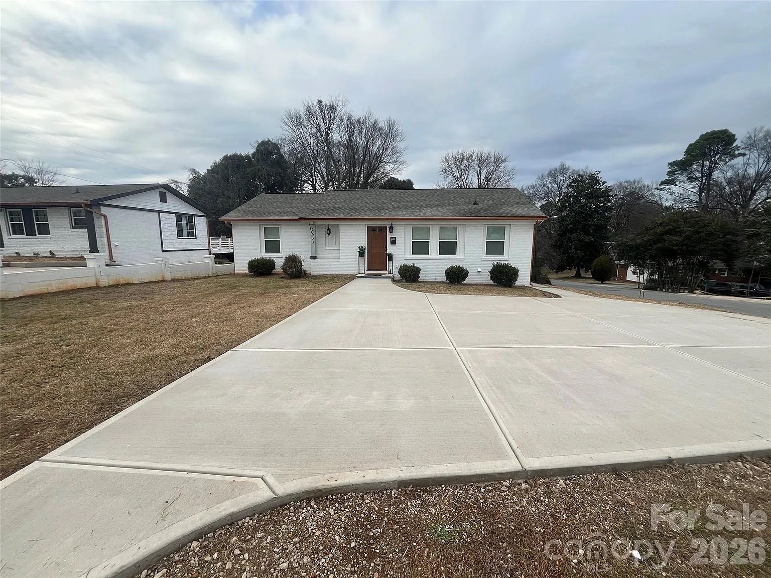 Single-story white brick house with brown roof, brown front door, and bushes in front. Large concrete driveway and brown grass lawn under cloudy sky.