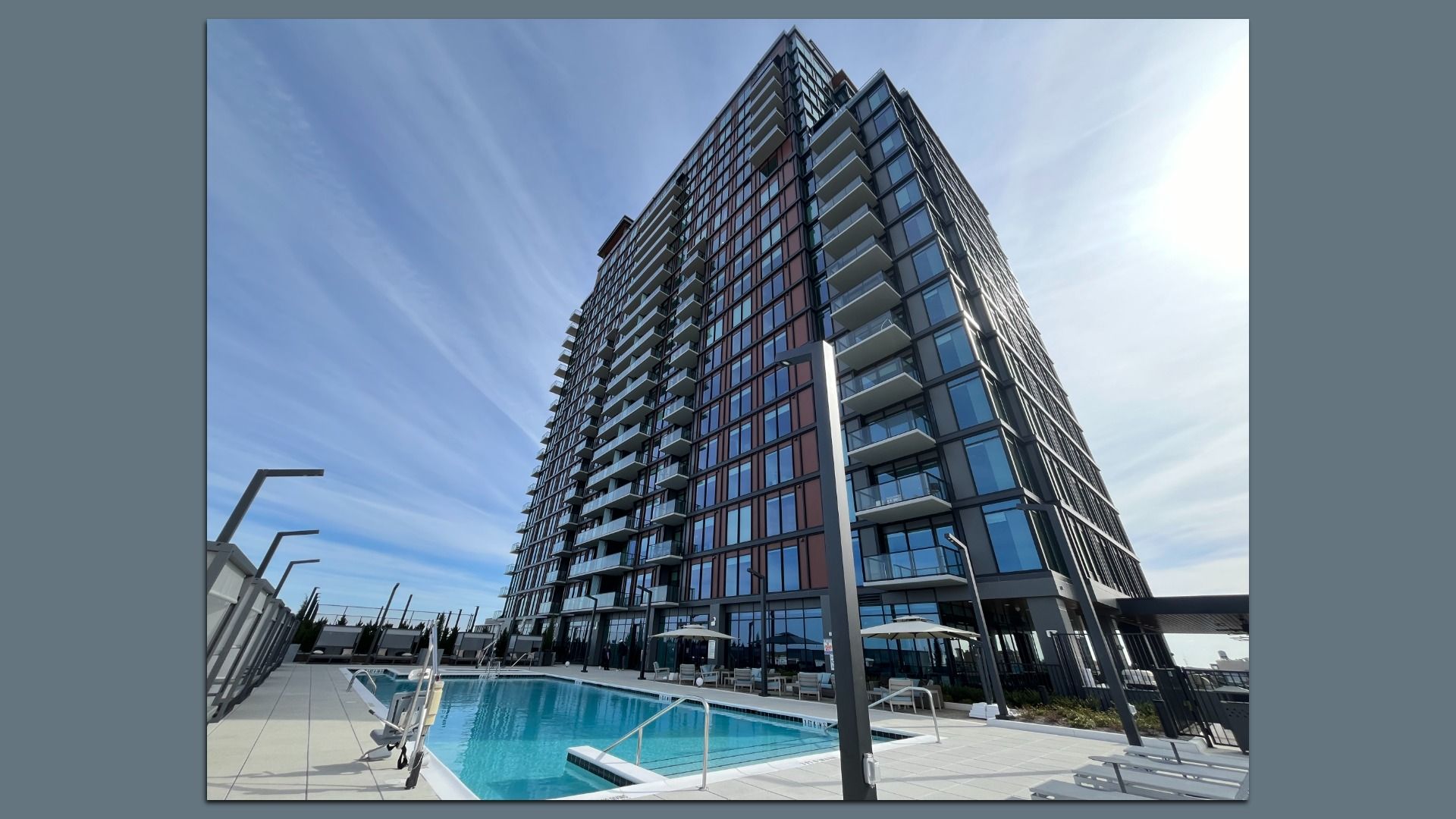 Tall glass-clad residential tower with numerous balconies looms over a pool deck; blue sky above, with lounge chairs and umbrellas around the pool.