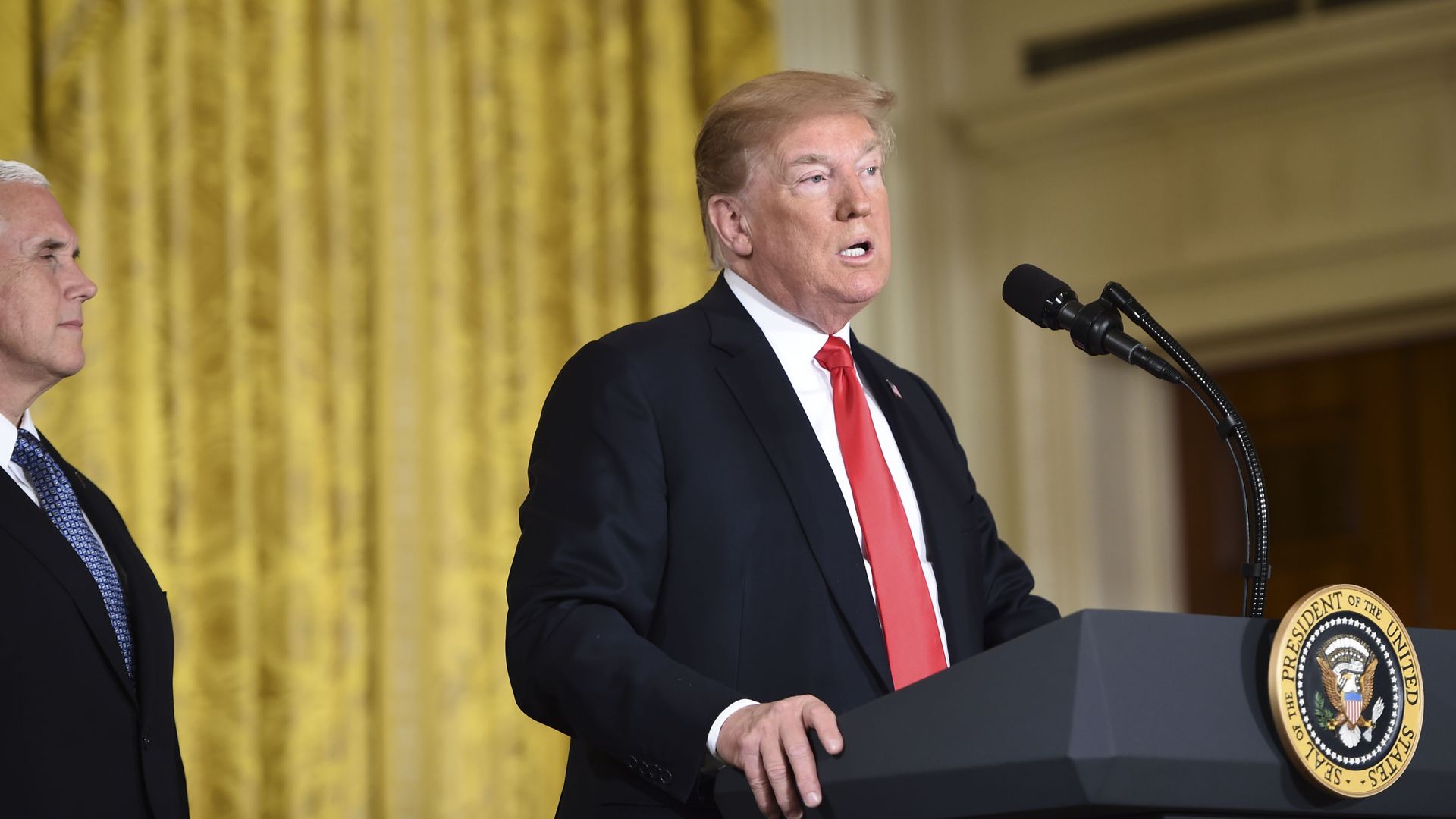 Donald Trump speaks at a podium before a gold drape.
