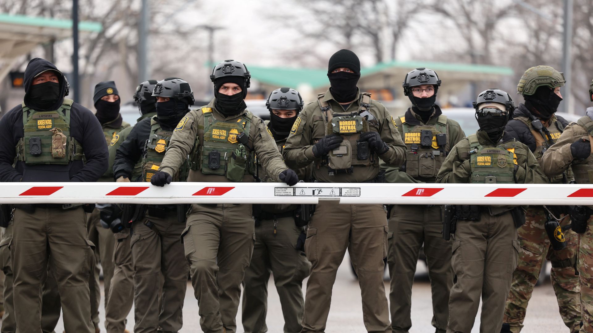 A large group of US Border Patrol agents in uniforms and masks stand behind a barrier outside