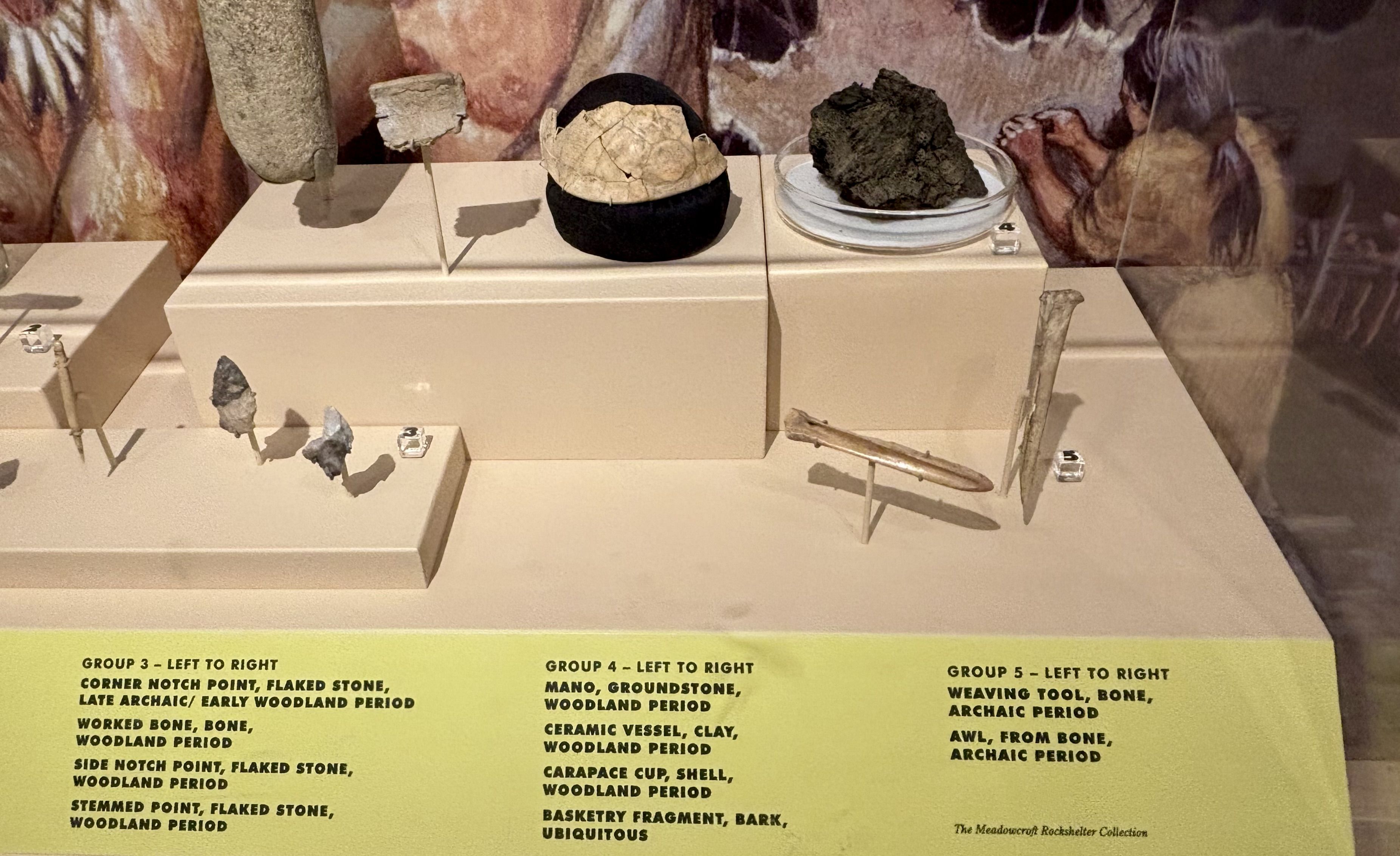Exhibit of ancient tools and pottery on beige pedestals: cracked ceramic vessel on a black base center, rough rock on a plate to the right, small flaked stones in front, with a yellow caption at the bottom.