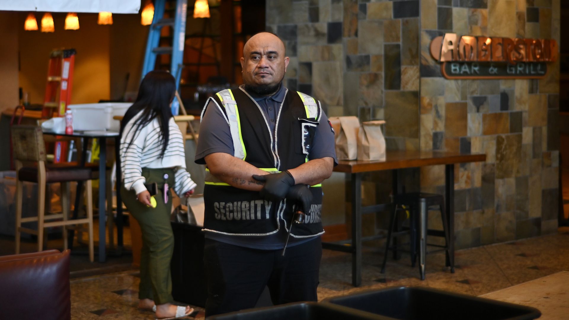 A man stands with his arms held together while wearing a short sleeve shirt and a light vest with the word SECURITY over the shirt. Behind him, a hotel lobby is visible, and a woman can be seen walking nearby.