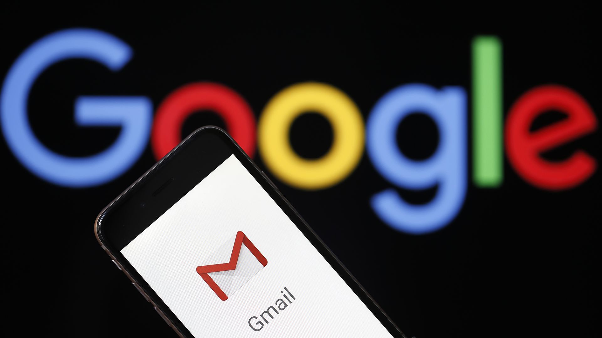 In this image, a phone screen with the Gmail logo is held in front of a large Google logo. 