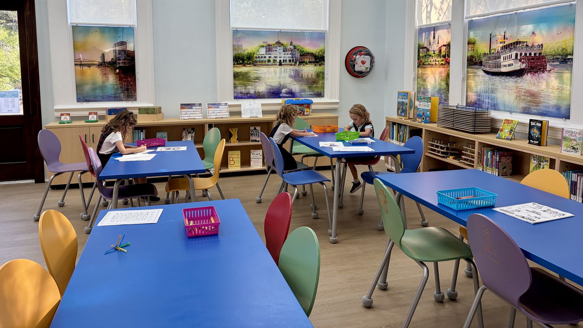 Bright classroom with blue tables and multicolored chairs. Children draw at desks as large window murals show boats and cityscapes; bookshelves line the right wall.