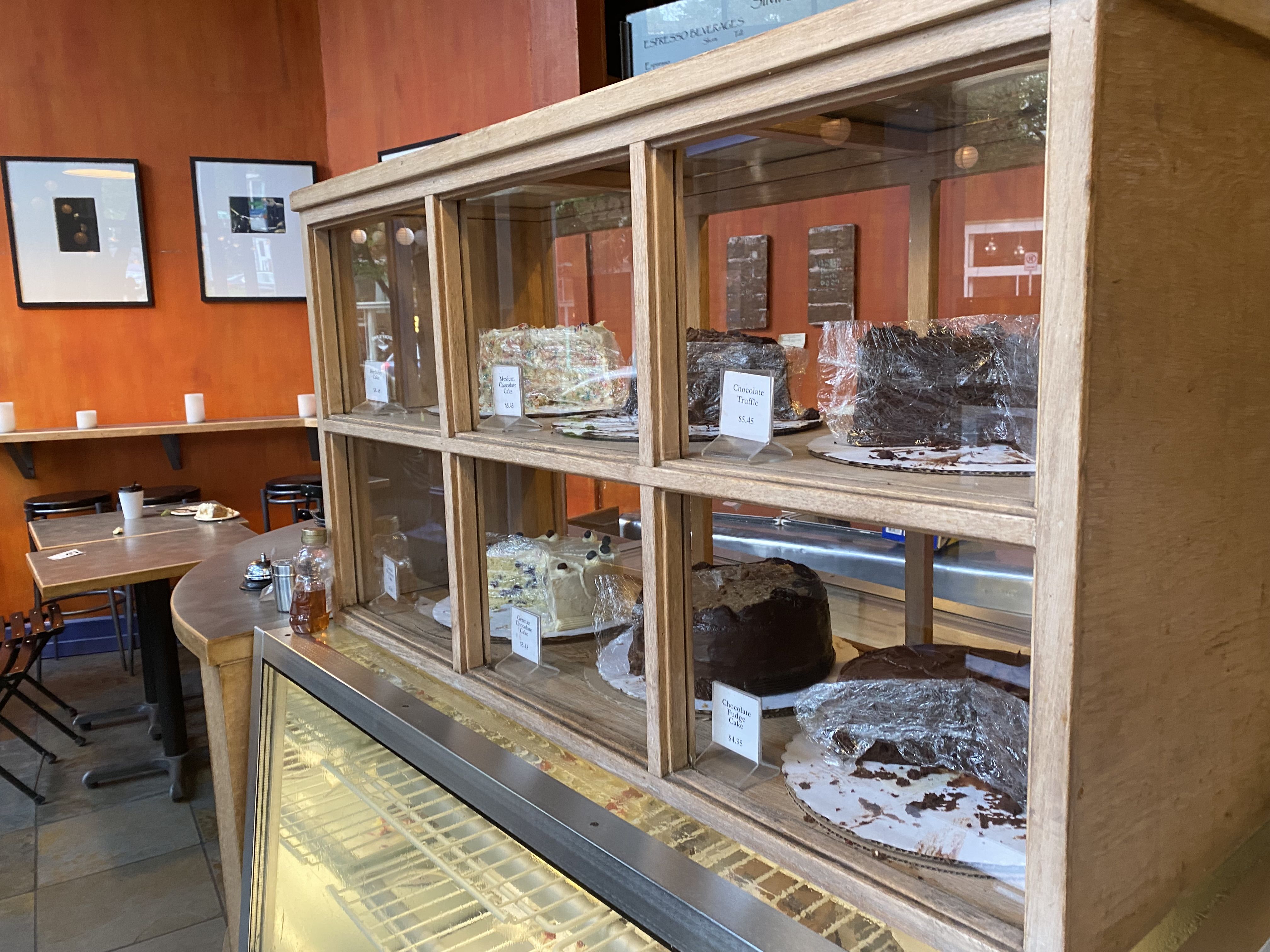 A view of a display case with six cakes, with two chocolate-based ones in the foreground, and a cafe area with tables in the background. 