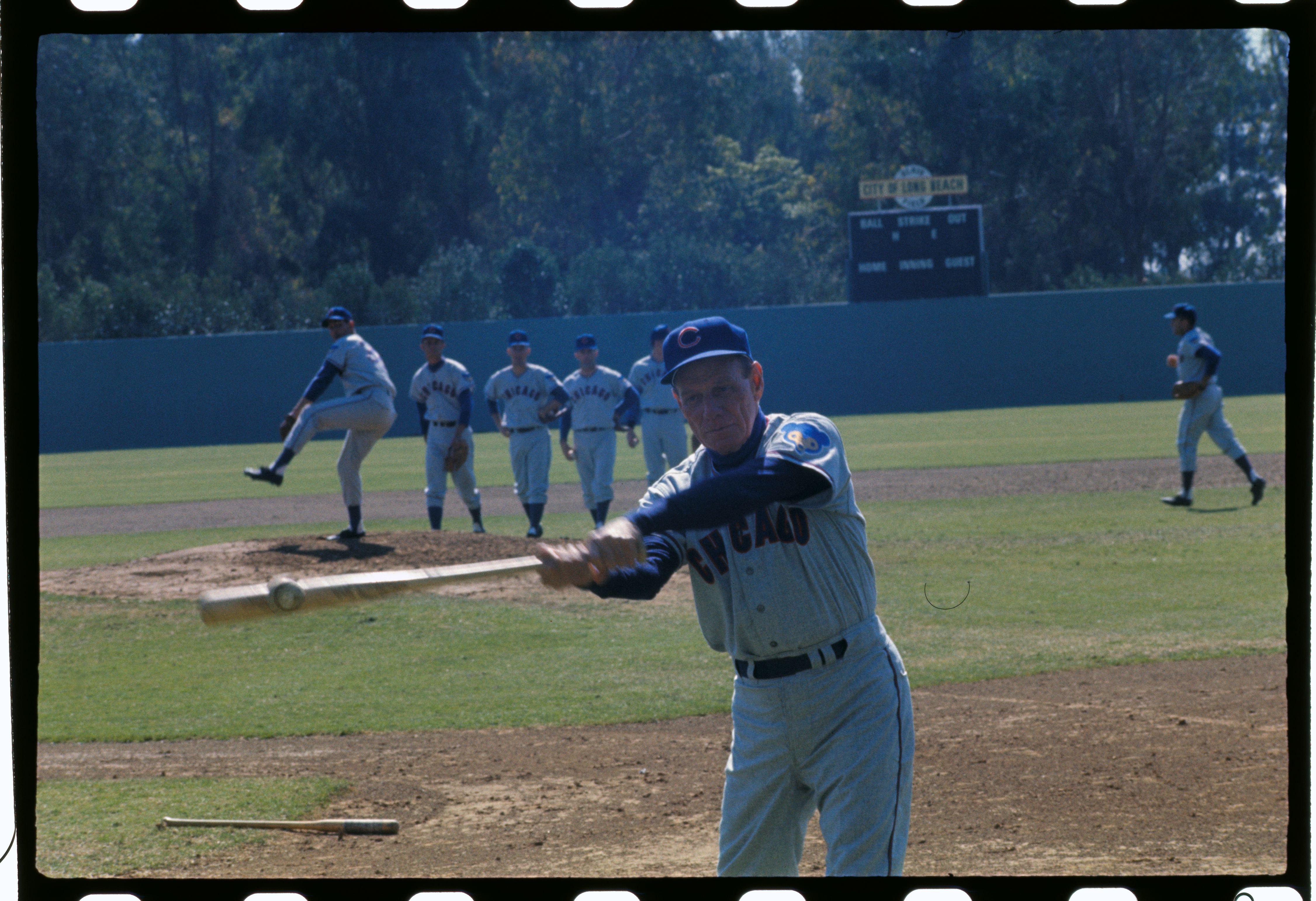Photo of a coach hitting a baseball with a bat while players warm up on the field. 