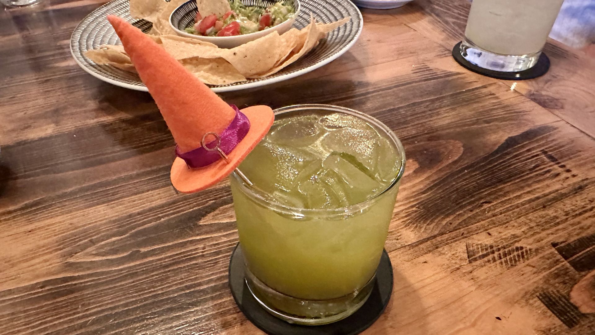 Yellow-green cocktail with ice in a glass on a wooden table, topped by a small orange hat with a purple ribbon. In the background, a bowl of chips and dip on a patterned plate.