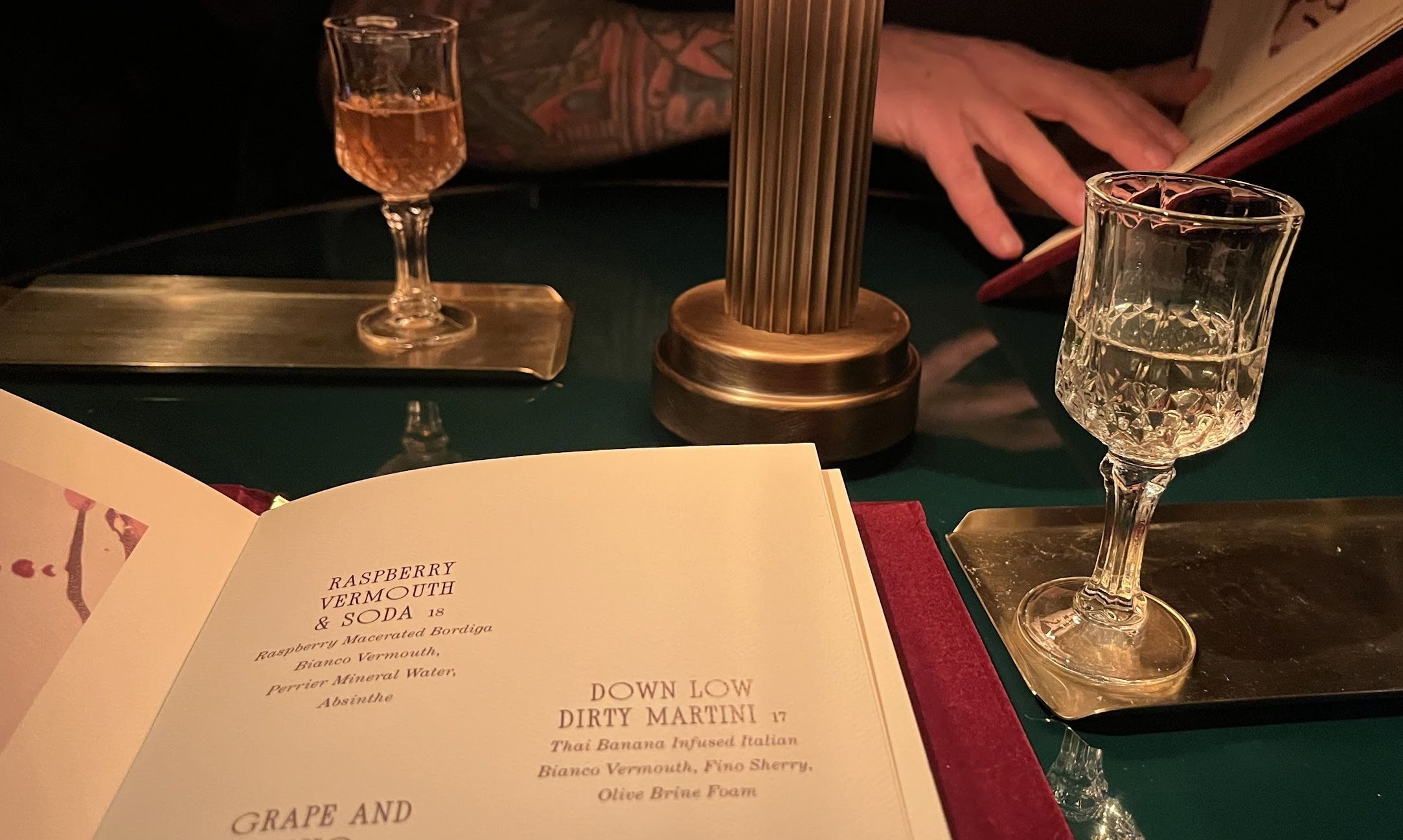 Open menu on a green bar with two crystal glasses, a brass lamp in the center, and a hand turning a page; drinks include "Raspberry Vermouth & Soda" and "Down Low Dirty Martini".