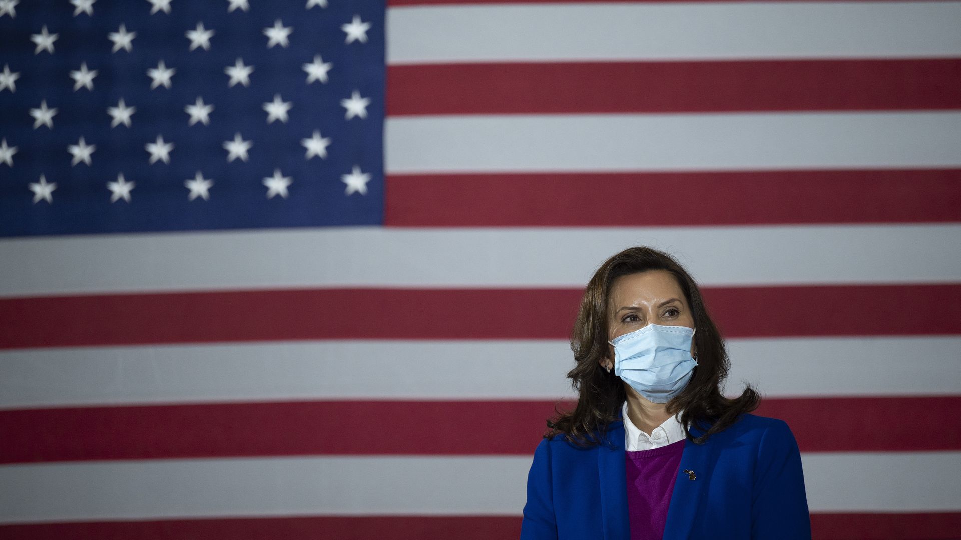 Photo of Gretchen Whitmer wearing a mask, with the American flag as a backdrop