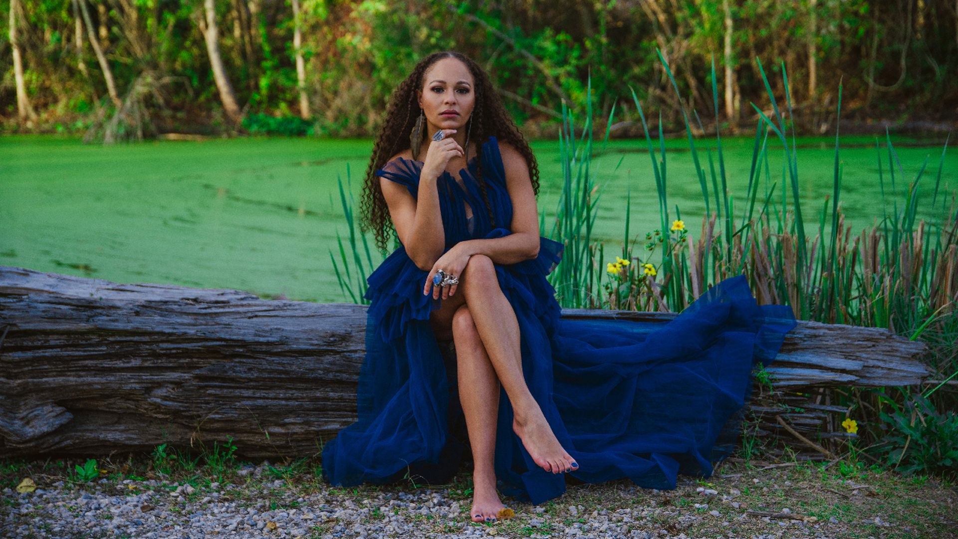 Woman with long curly hair in a flowing dark blue dress sitting barefoot on a large log by a green pond with tall grass and yellow flowers, surrounded by trees.