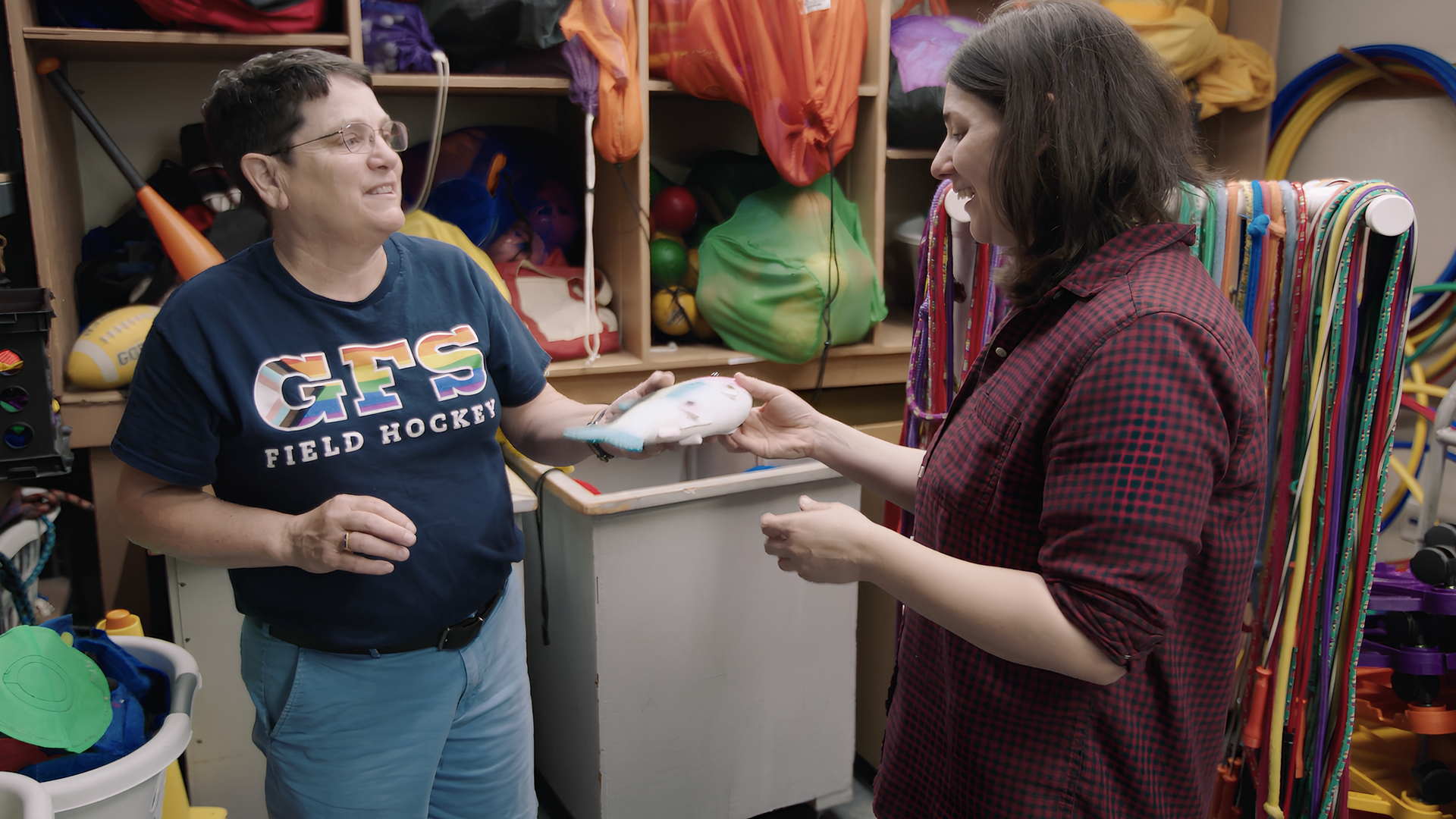 Two adults in a colorful, cluttered playroom exchange a white item with blue trim; shelves behind them hold toys, ribbons, and sports gear.