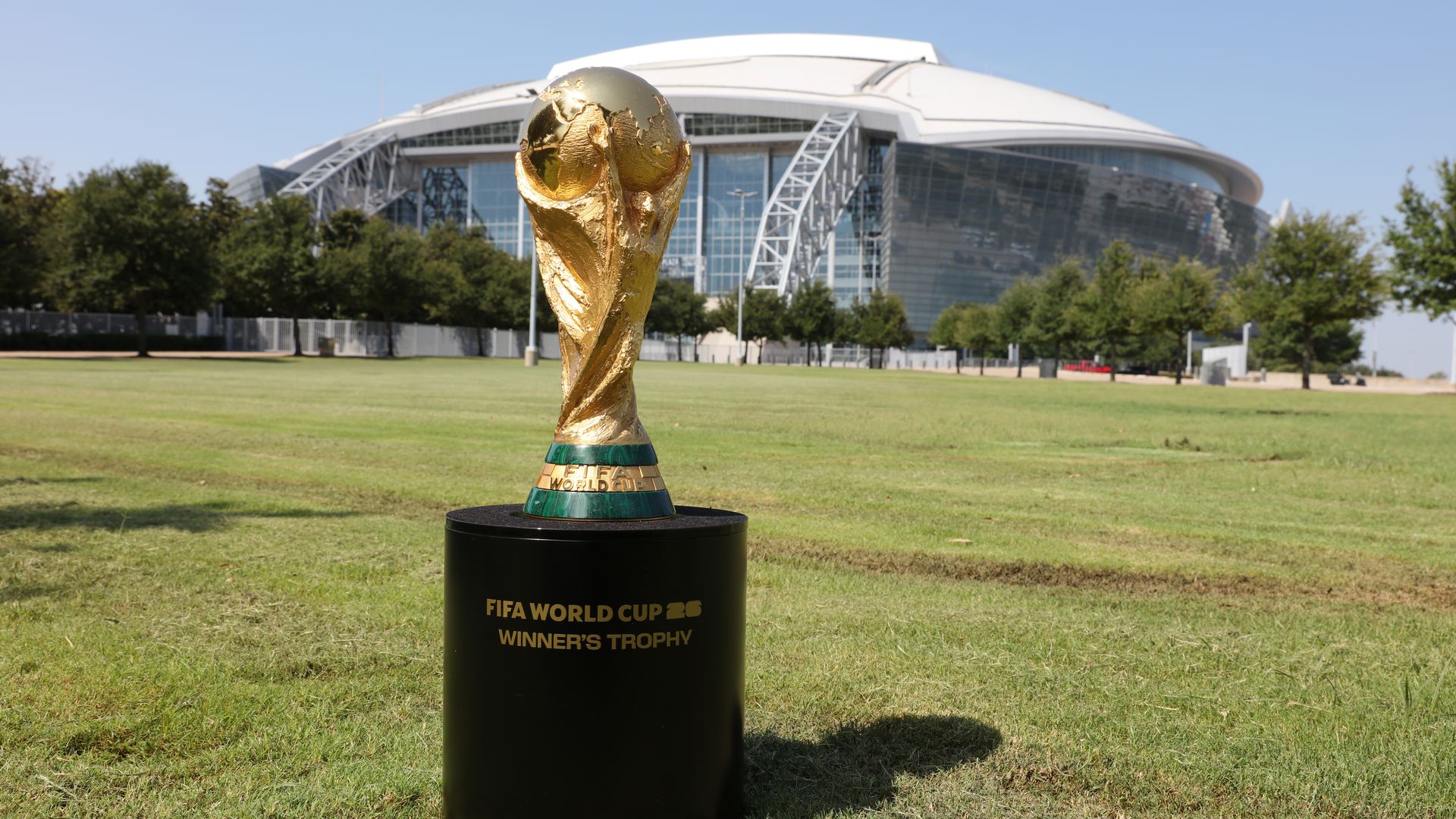 The World Cup trophy on a lawn outside AT&T Stadium, with the stadium in the background 