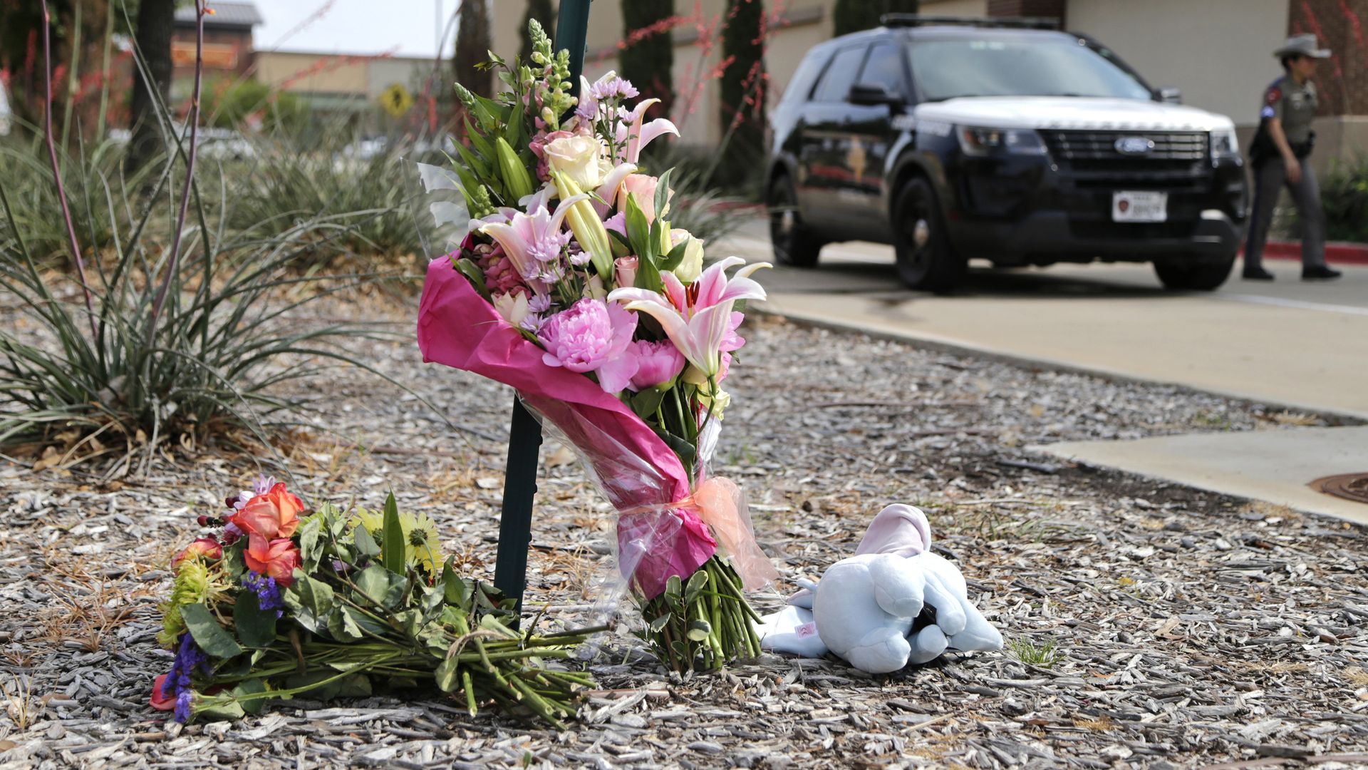 Two bouquets of flowers and a stuffed animal on the ground in front of a police car at the Allen outlet mall
