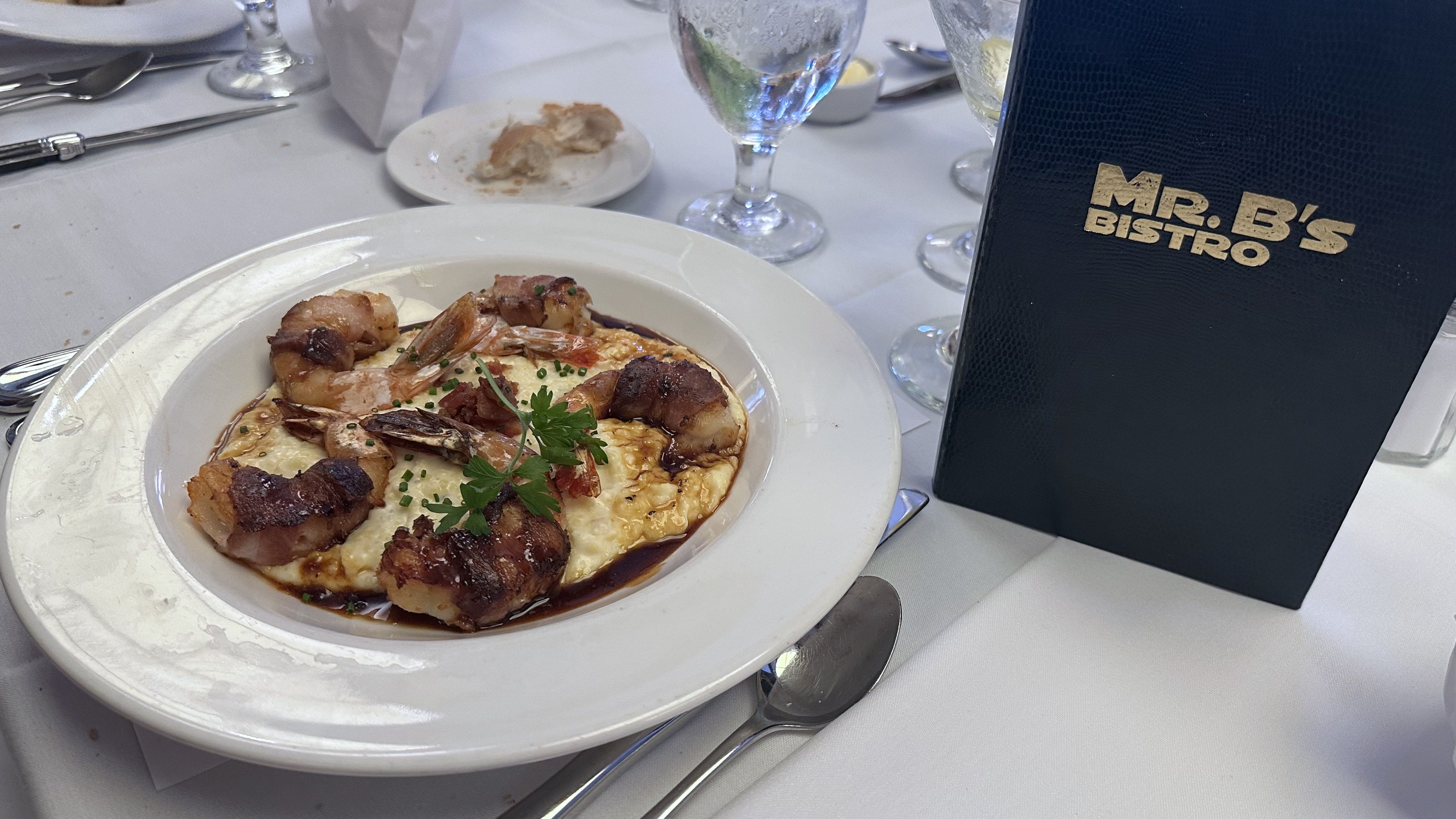 White plate with shrimp wrapped in bacon on creamy grits, garnished with parsley and chives, on a white tablecloth with a water glass and a navy menu labeled "Mr. B's Bistro".