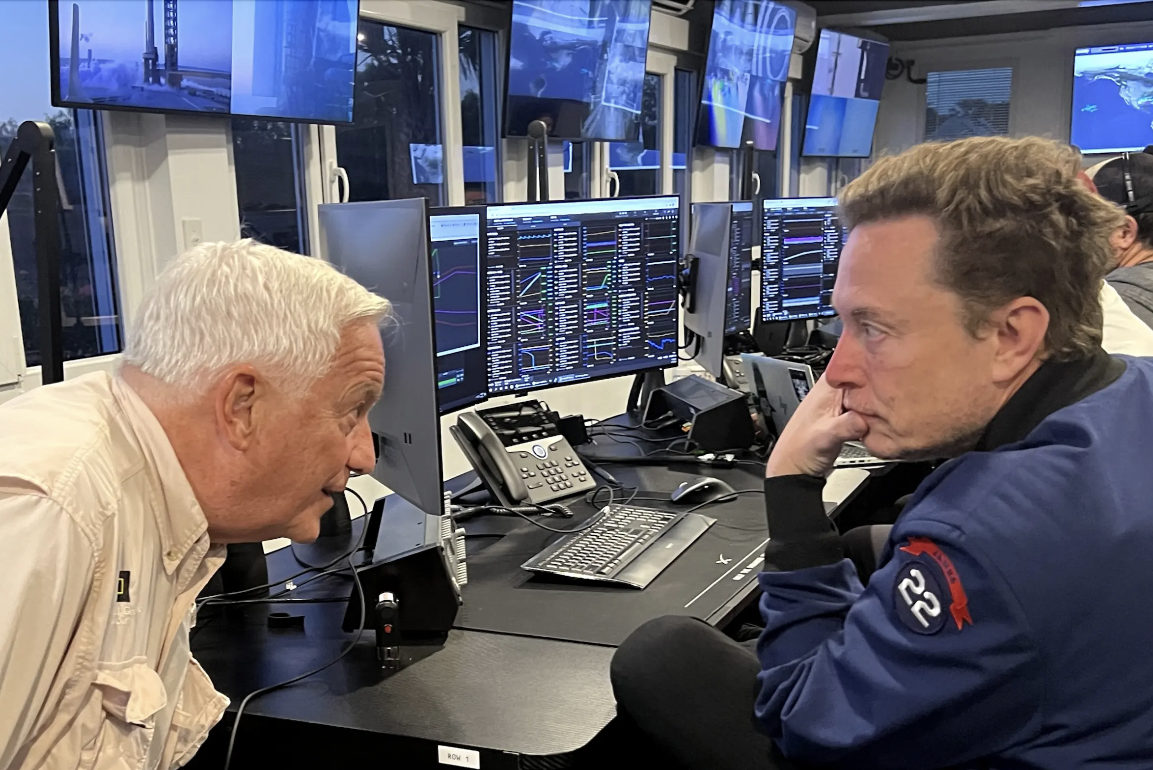 Walter Isaacson leans over to speak with Elon Musk in front of computer terminals