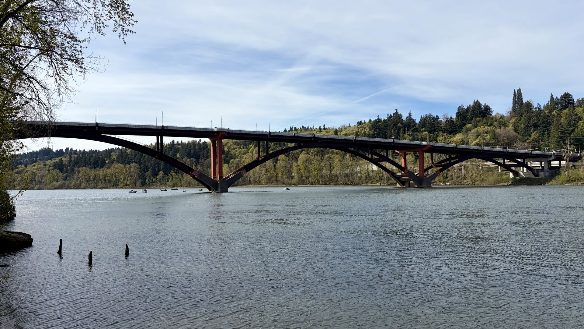An arched bridge with red supports spans a calm river, flanked by forested hills. A few boats glide on the water; the left foreground shows tree branches framing the scene under a blue sky.