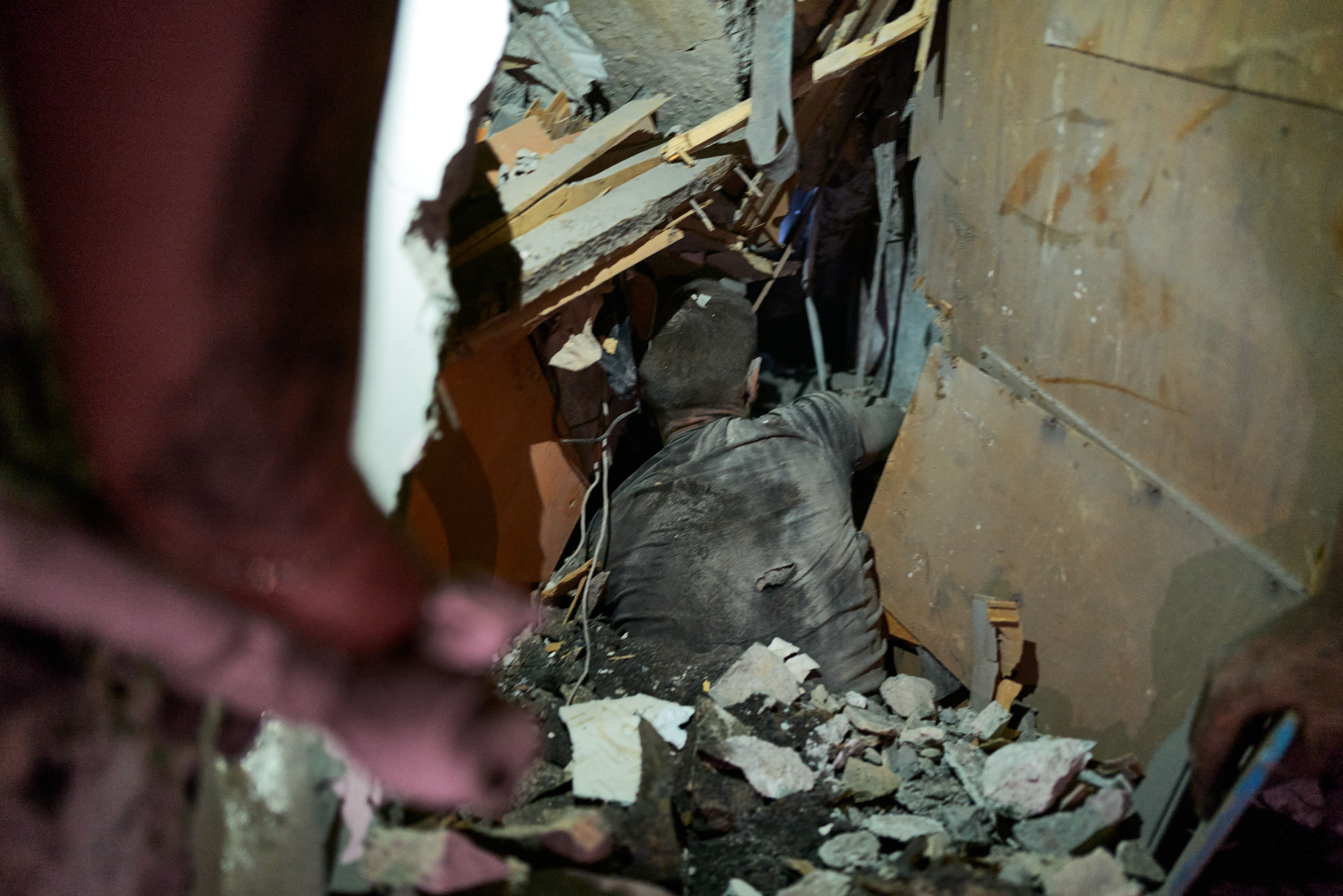 A person covered in dust is crawling through a narrow rubble-filled space with broken wood, concrete, and metal debris around them, suggesting a building collapse or disaster site.