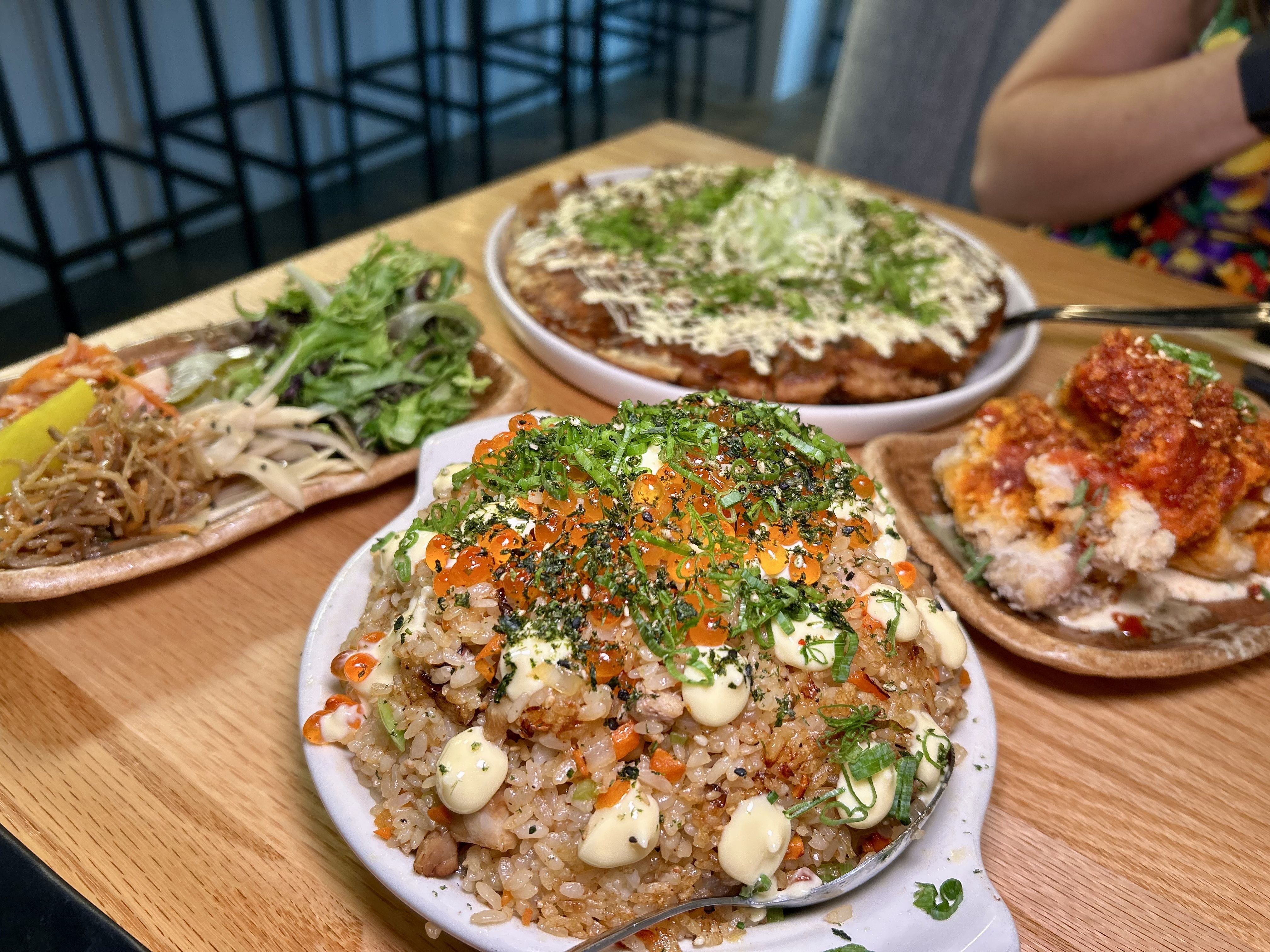 A table setting with several dishes of Asian cuisine. In the foreground is a plate of fried rice garnished with mayonnaise, green onions, and fish roe. To the left, there's a plate of mixed greens, pickled vegetables, and noodles.