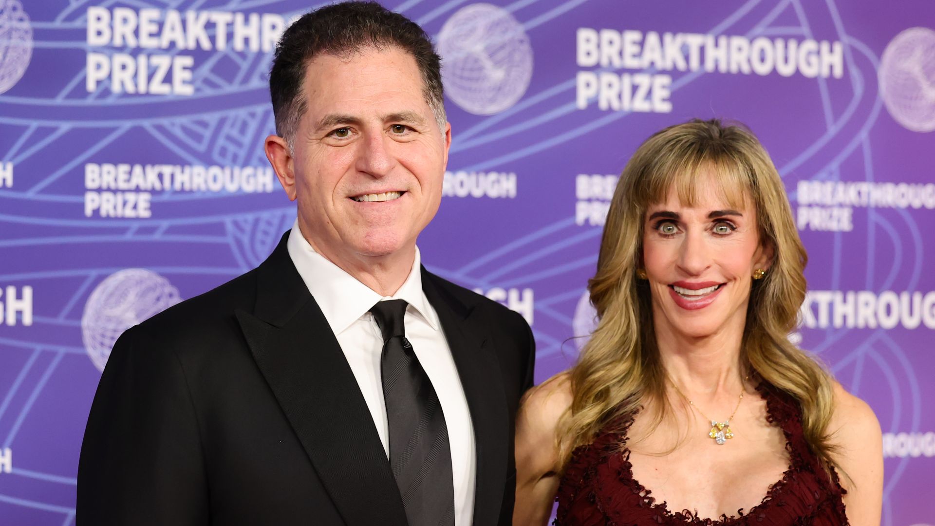 A man in a black suit and tie stands beside a smiling woman in a burgundy gown on a purple backdrop patterned with "BREAKTHROUGH PRIZE" text.