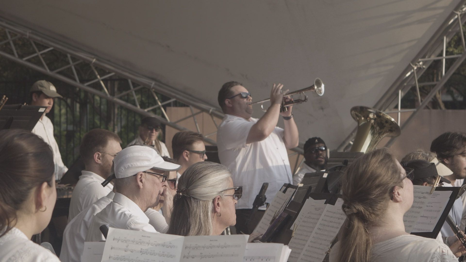 Outdoor brass band on stage; a trumpeter in a white shirt and sunglasses plays center, surrounded by musicians with sheet music; a tuba is visible on the right, audience in the foreground.