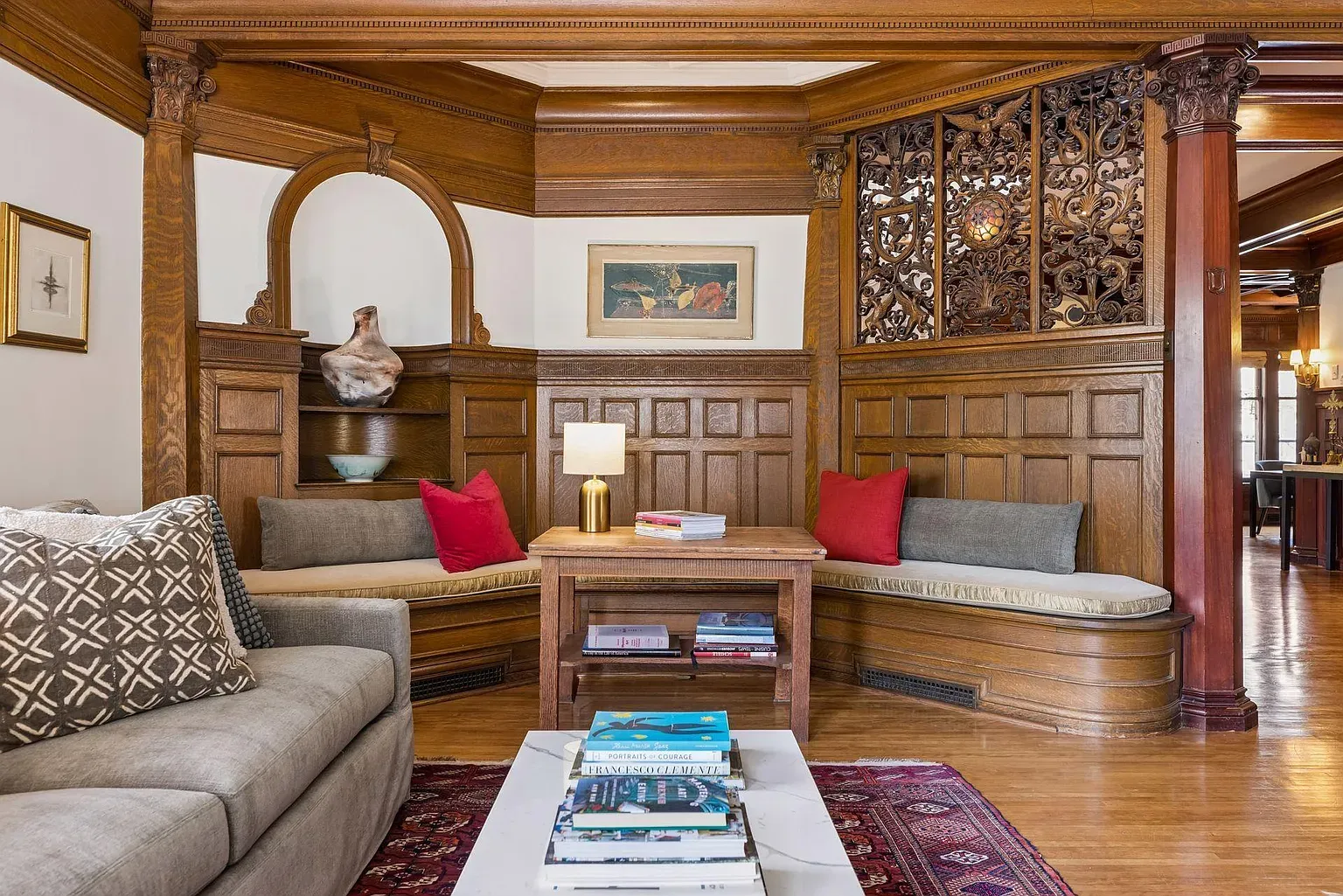 A traditional living room with dark wood paneling, curved bench seating with gray cushions and red pillows, a wooden coffee table, a patterned rug, decorative carved screens, and framed art.
