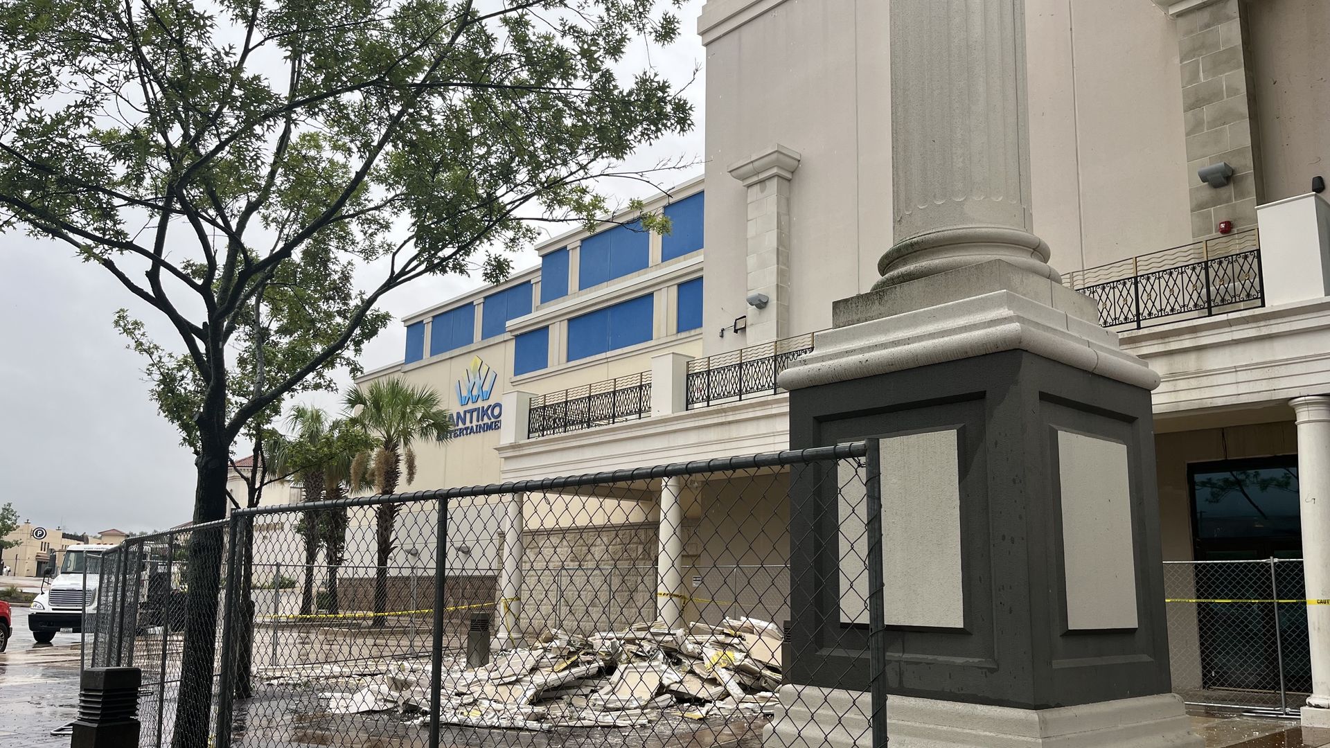 Street view of a beige neoclassical building with large pillars and a black metal railing; a chain-link fence, puddled wet pavement, a tree on the left, and scattered construction debris in front.
