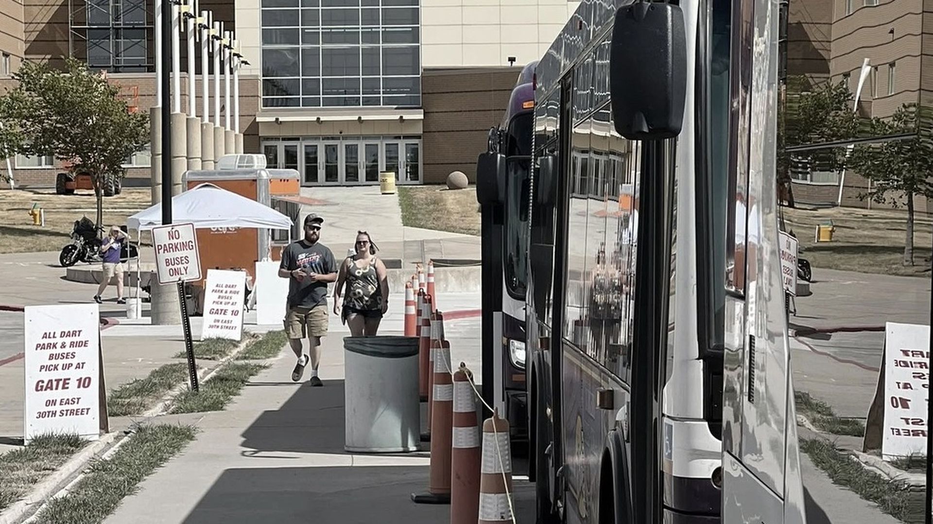 Campus bus pickup area with a silver bus on the right, orange cones, a white canopy, and a couple walking toward a glass-front building; signs read "Gate 10" and "Park & Ride".