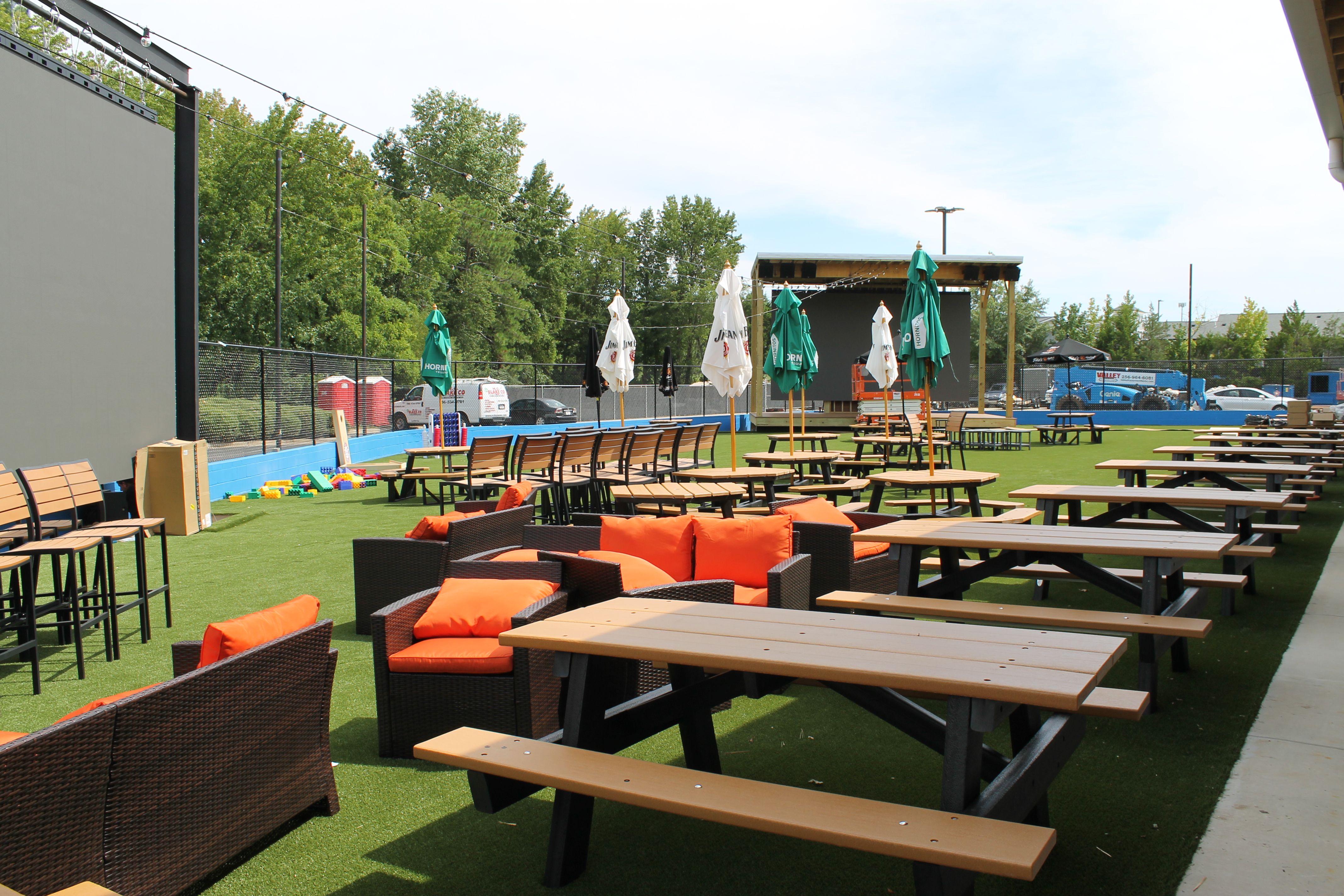 Outdoor seating area with brown picnic tables, wicker couches with bright orange cushions, green and white umbrellas, artificial grass, and a large screen on a wooden stage.