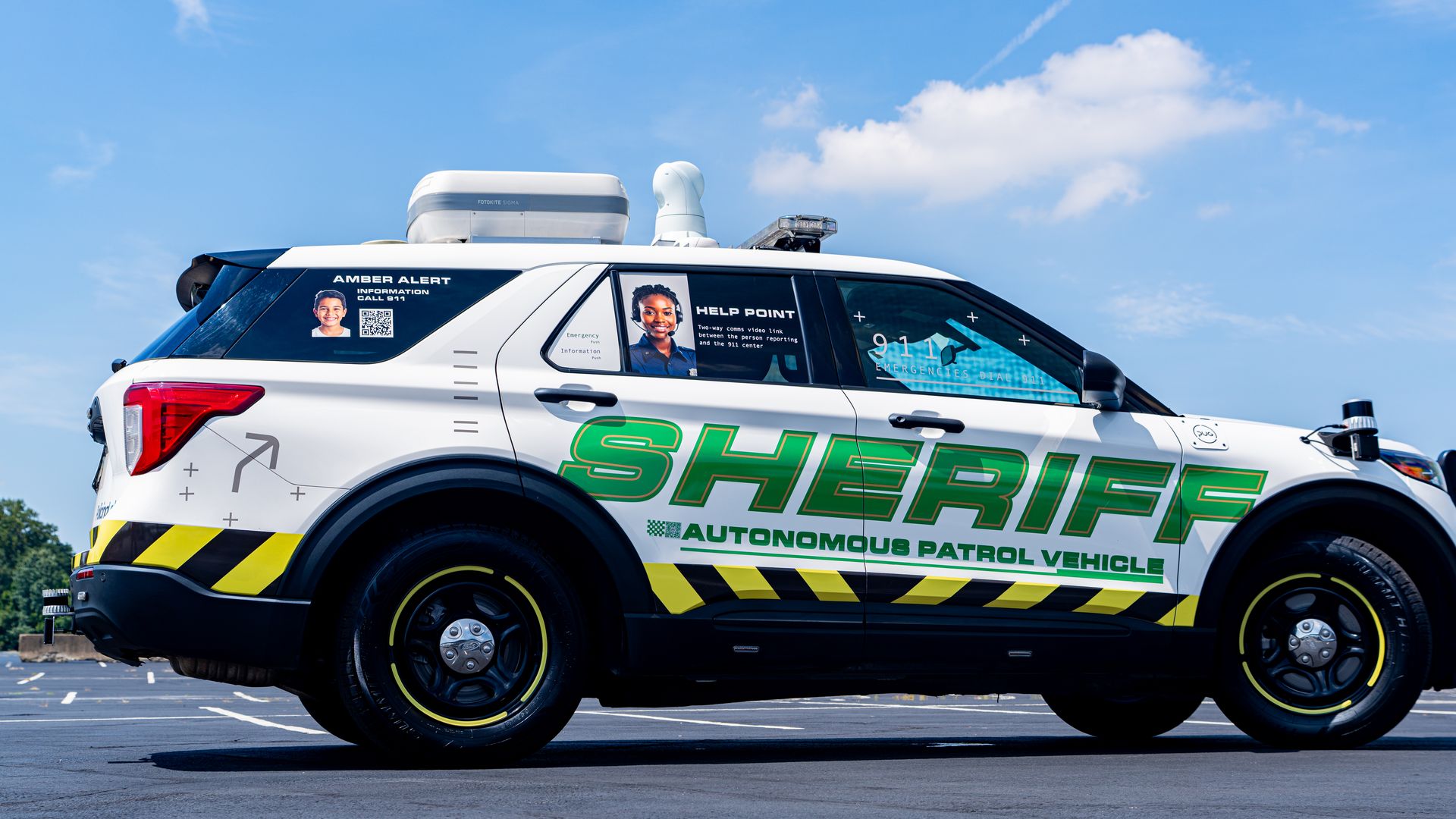White sheriff autonomous patrol vehicle with green and yellow markings, amber alert photo, and help point video link signage parked outdoors under a blue sky with clouds.