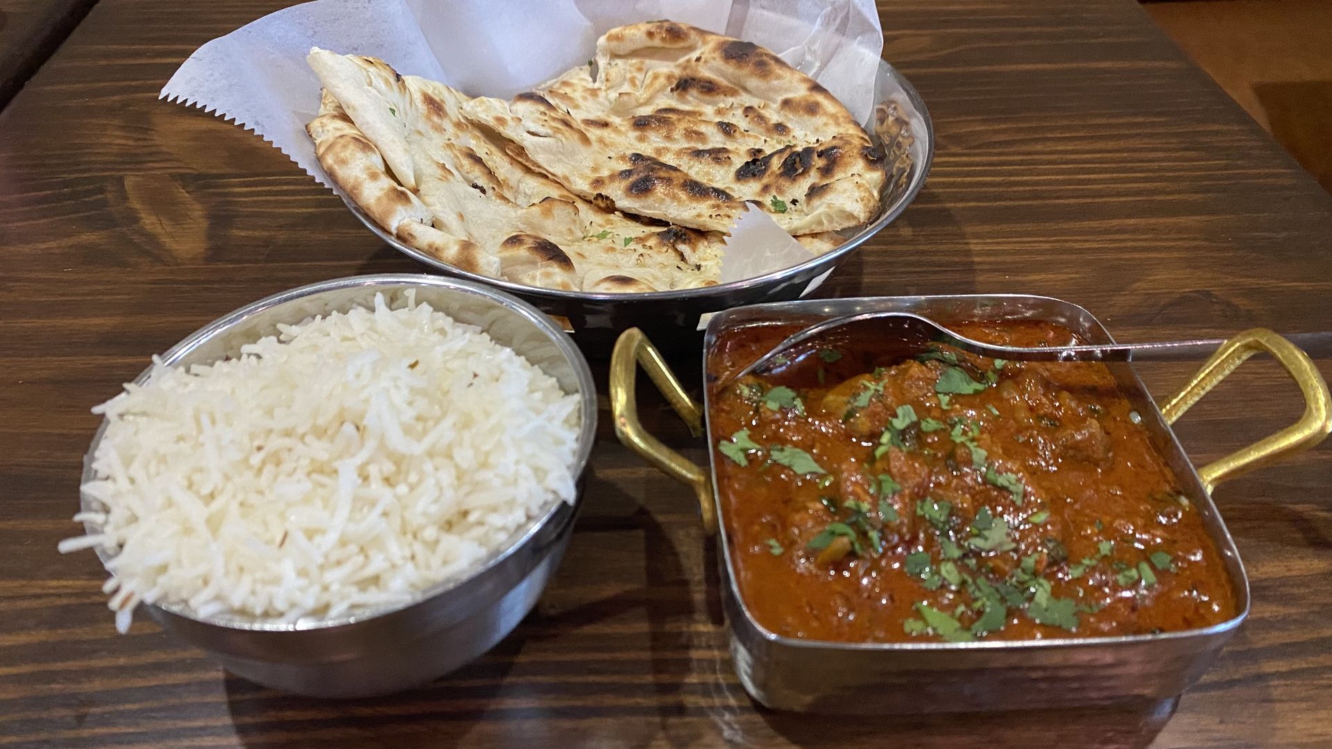 A bowl of Basmati rice, a bowl of sliced pieces of naan bread, and a square dish of reddish-brown curry on a table. 