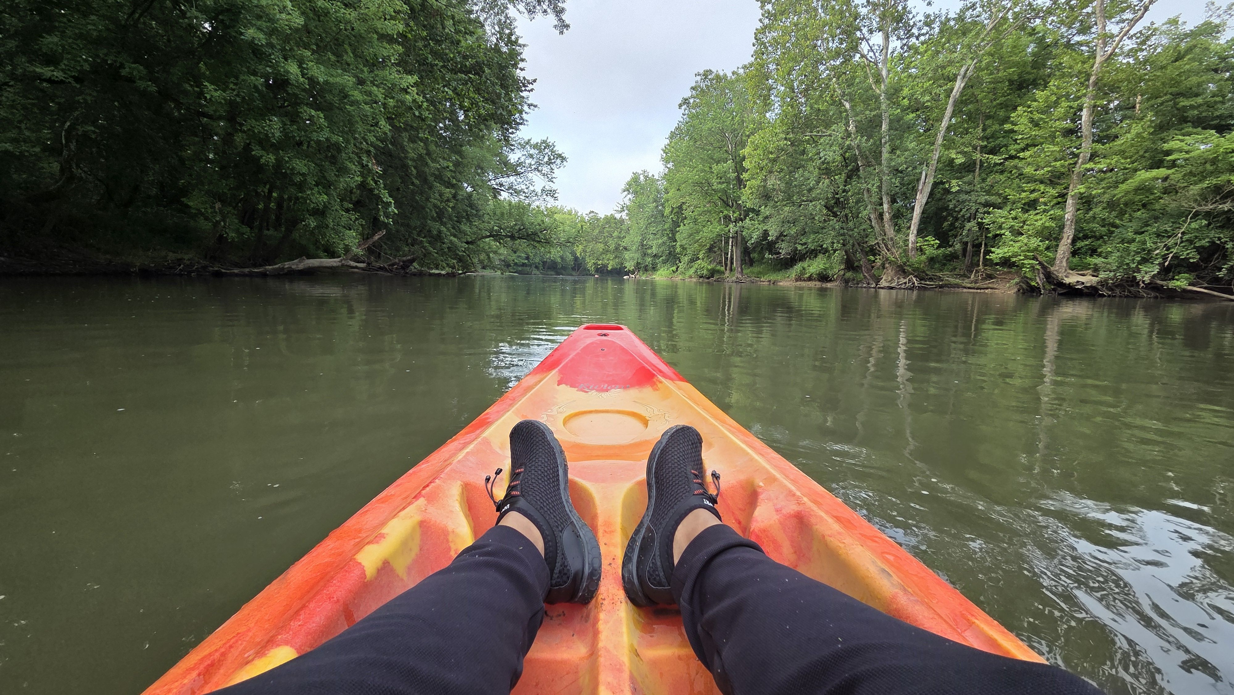 View from a kayak with red and yellow hues on a calm river surrounded by dense green trees, showing a person wearing black shoes and pants relaxing with feet stretched out.