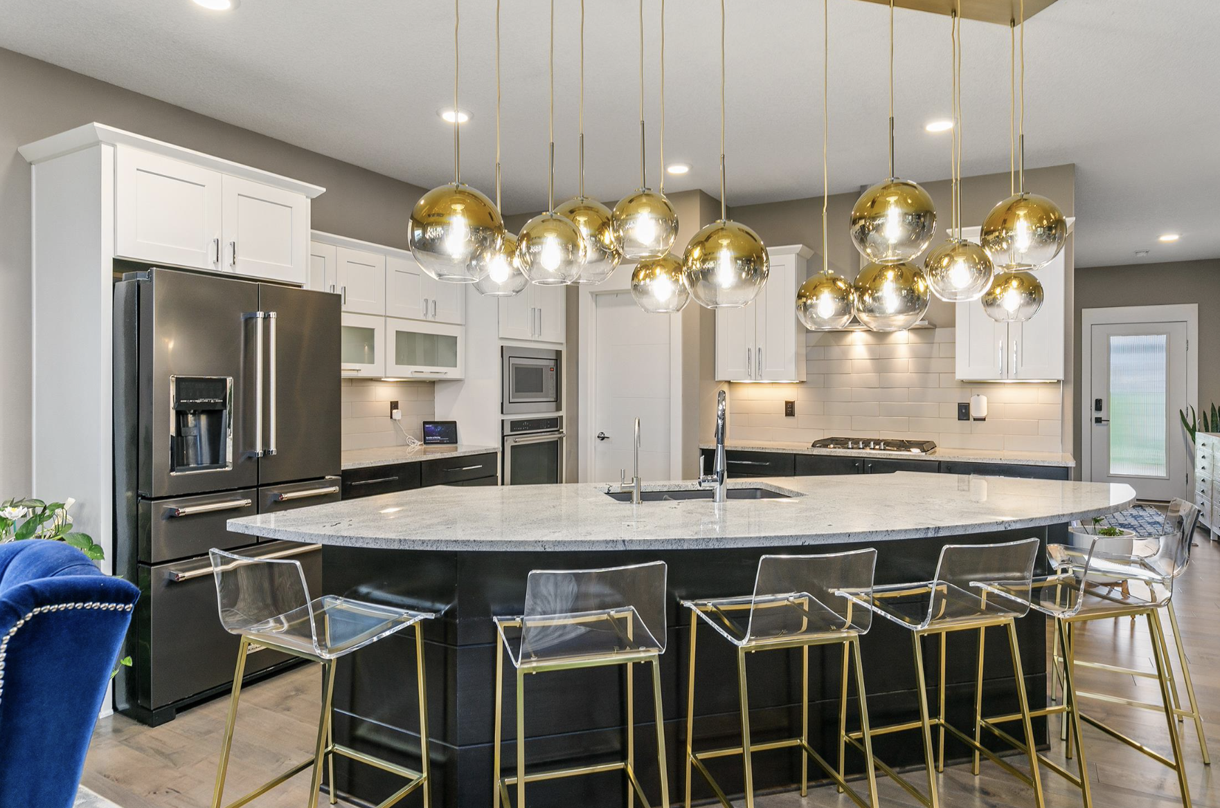 Modern kitchen with a large curved marble island, six transparent bar stools with gold legs, white cabinets, black refrigerator, and multiple gold spherical pendant lights hanging above.