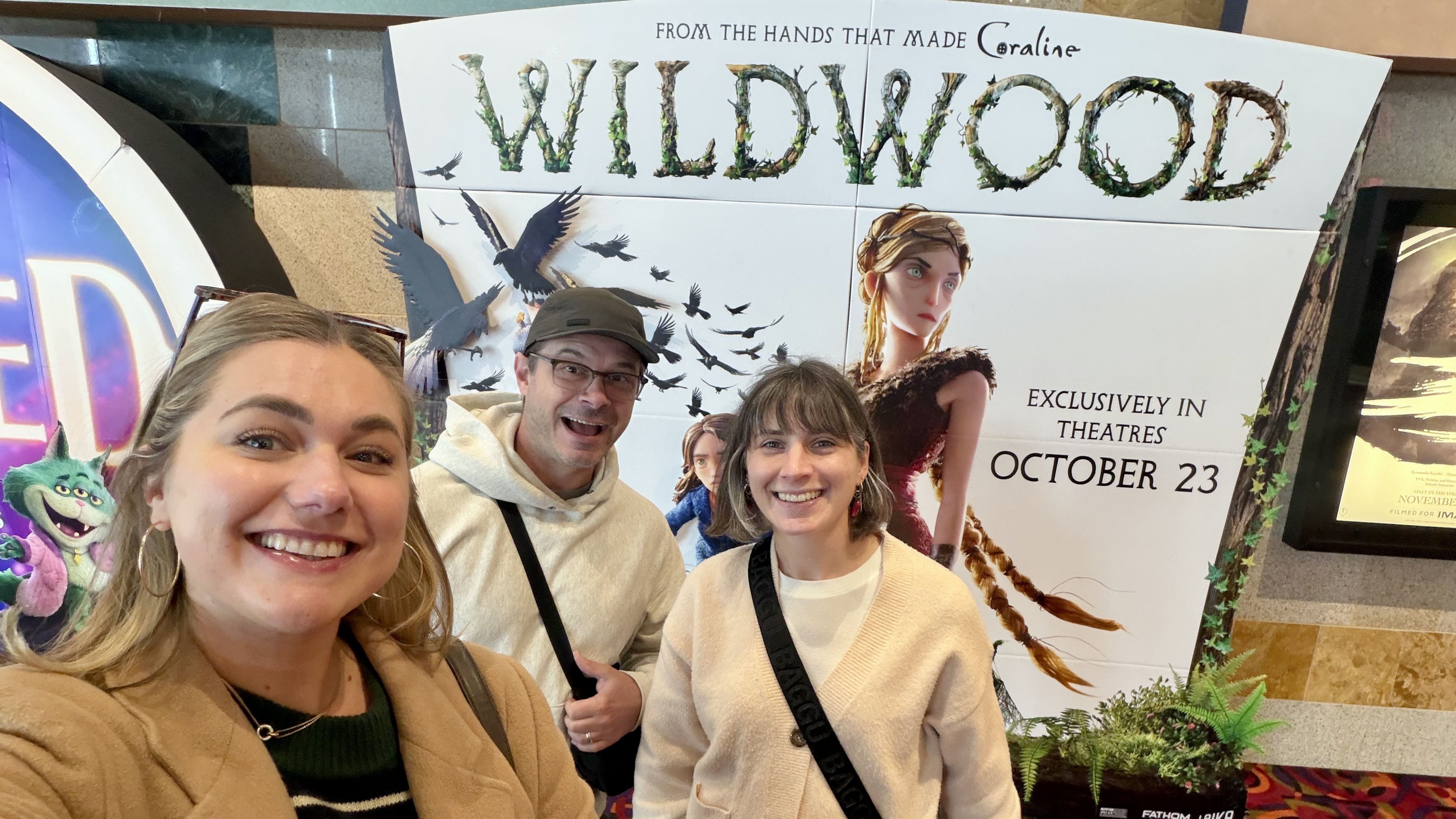 Three people take a selfie in front of a large poster for "WILDWOOD" with vine-covered letters, birds in flight, and a female character; text reads EXCLUSIVELY IN THEATRES OCTOBER 23.
