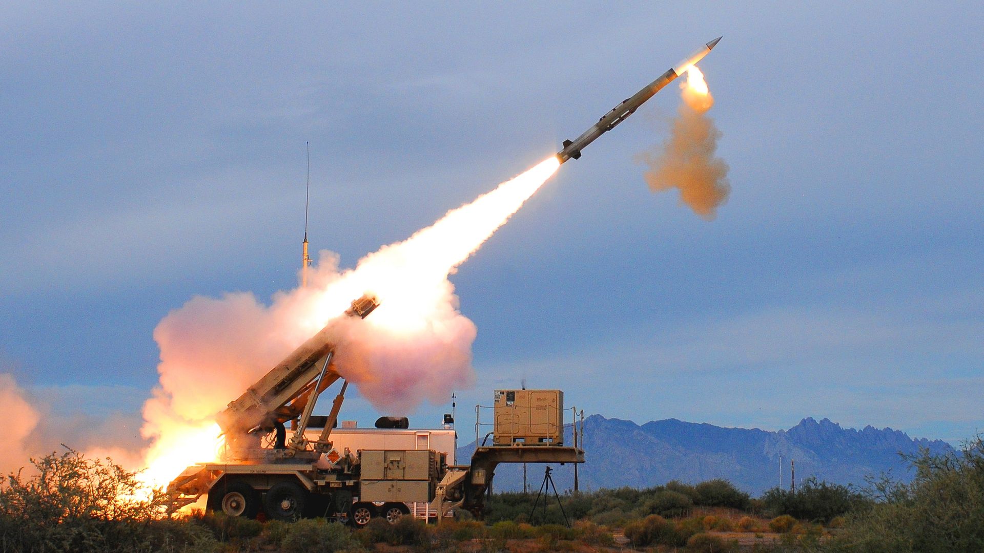Military missile launcher on a trailer amid desert scrub launches a missile with a bright flame and smoke trail against a cloudy blue sky and distant mountains.