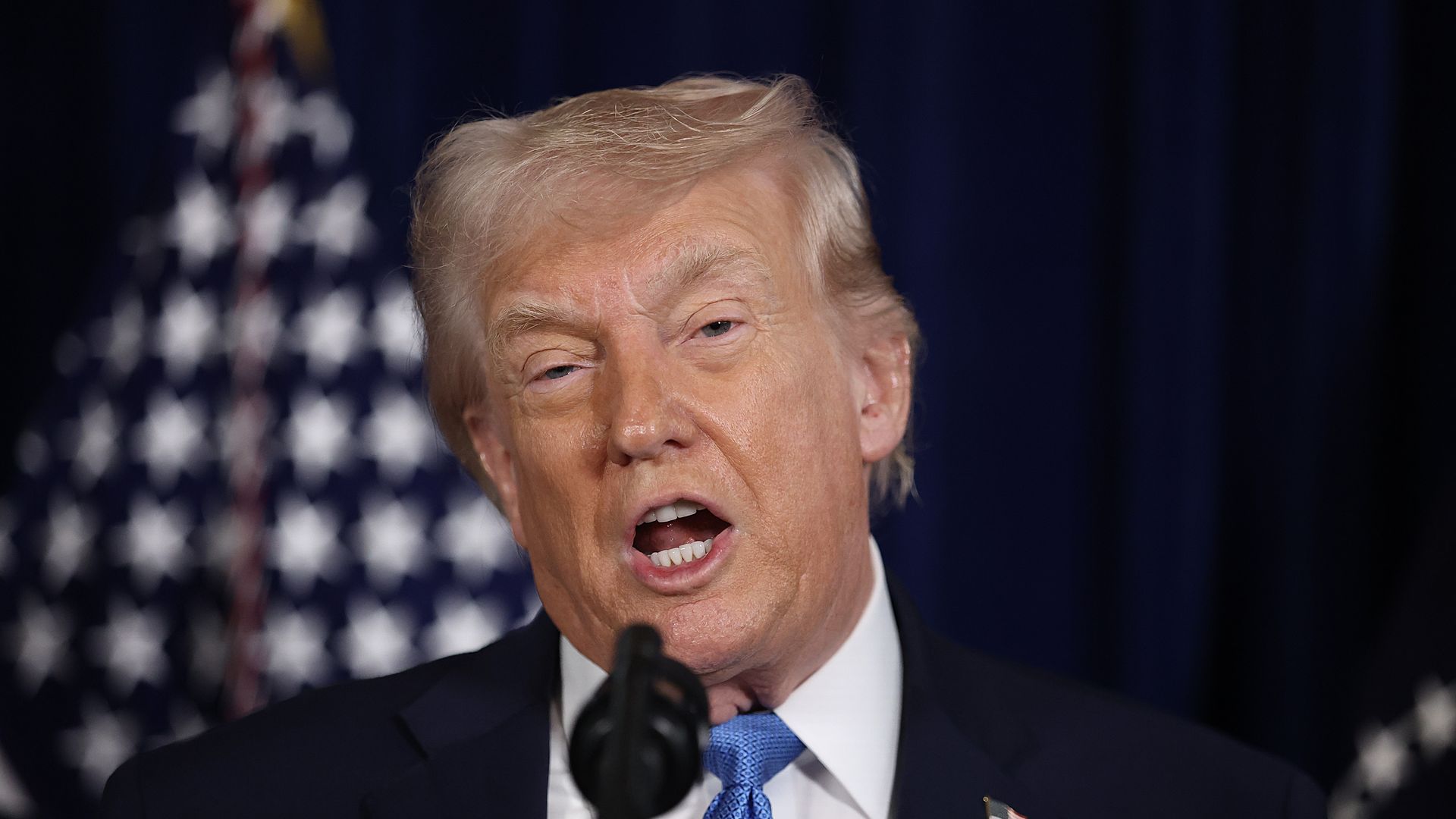 Trump speaking into a microphone wearing a navy suit jacket, white collared shirt and blue tie